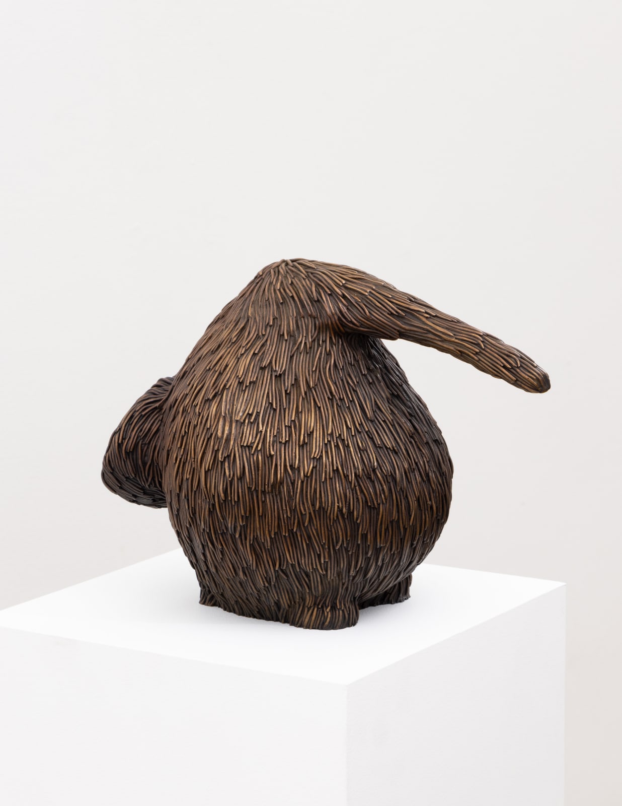 Free-standing bronze sculpture of a form resembling a kiwi with a long beak. The sculpture is cast with a surface texture resembling shaggy hair and treated with a golden brown patina. 