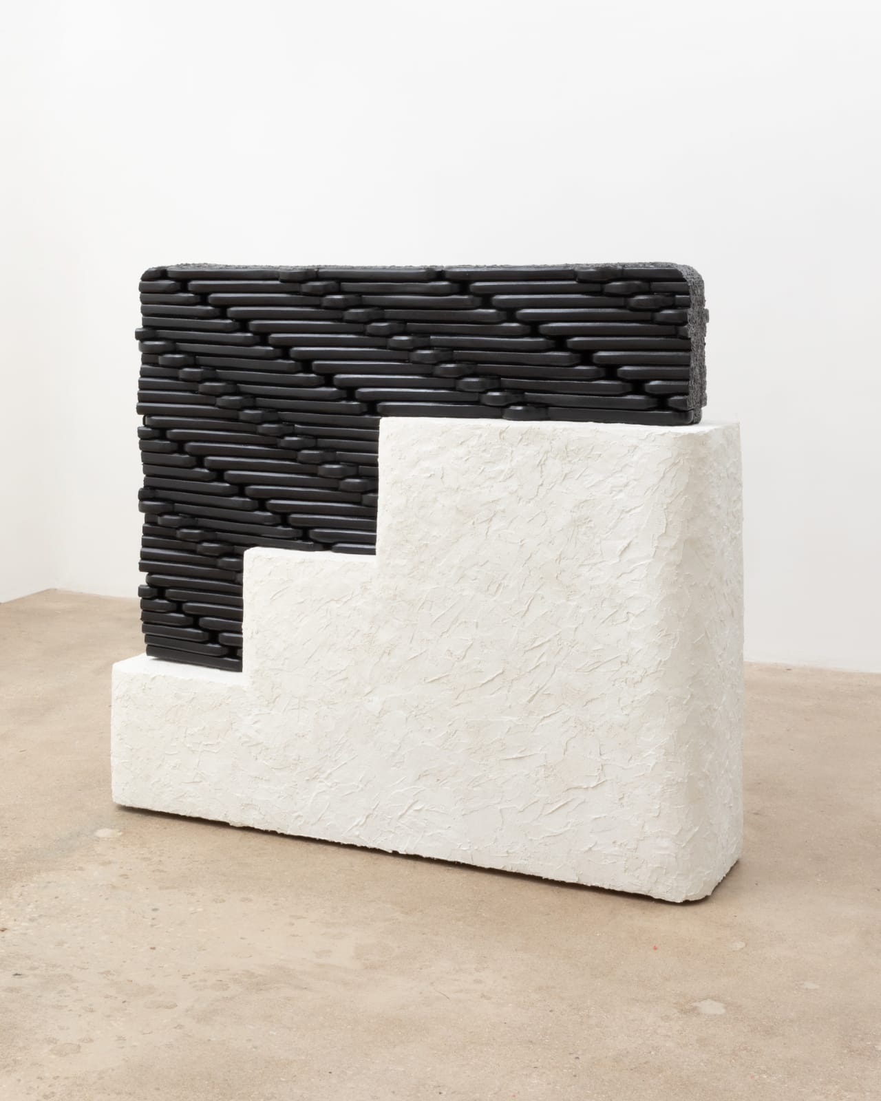 Free-standing sculpture consisting of carved polystyrene pieces aggregated together to form a black hued, interlocking patterned form that fits like a puzzle onto a white rectangular stucco base with tiered steps like a staircase.