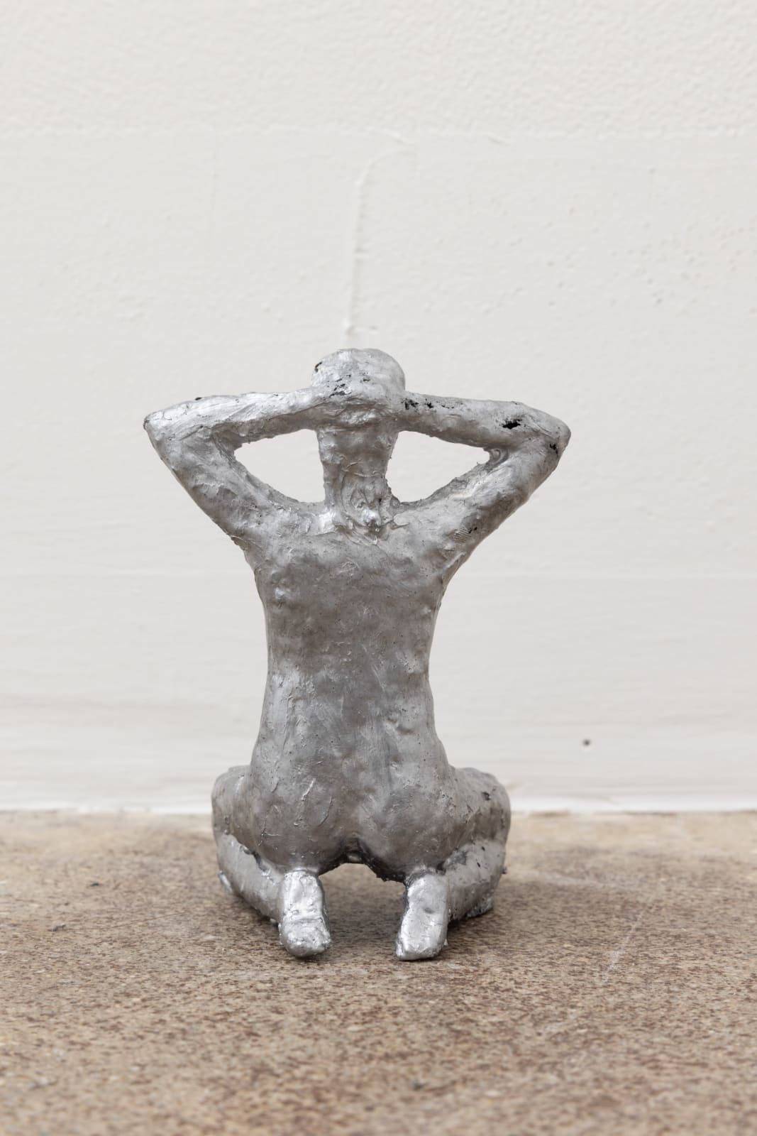Free-standing sculpture made from lost-wax cast aluminum featuring a kneeling human figure with hands on the back of the head. 
