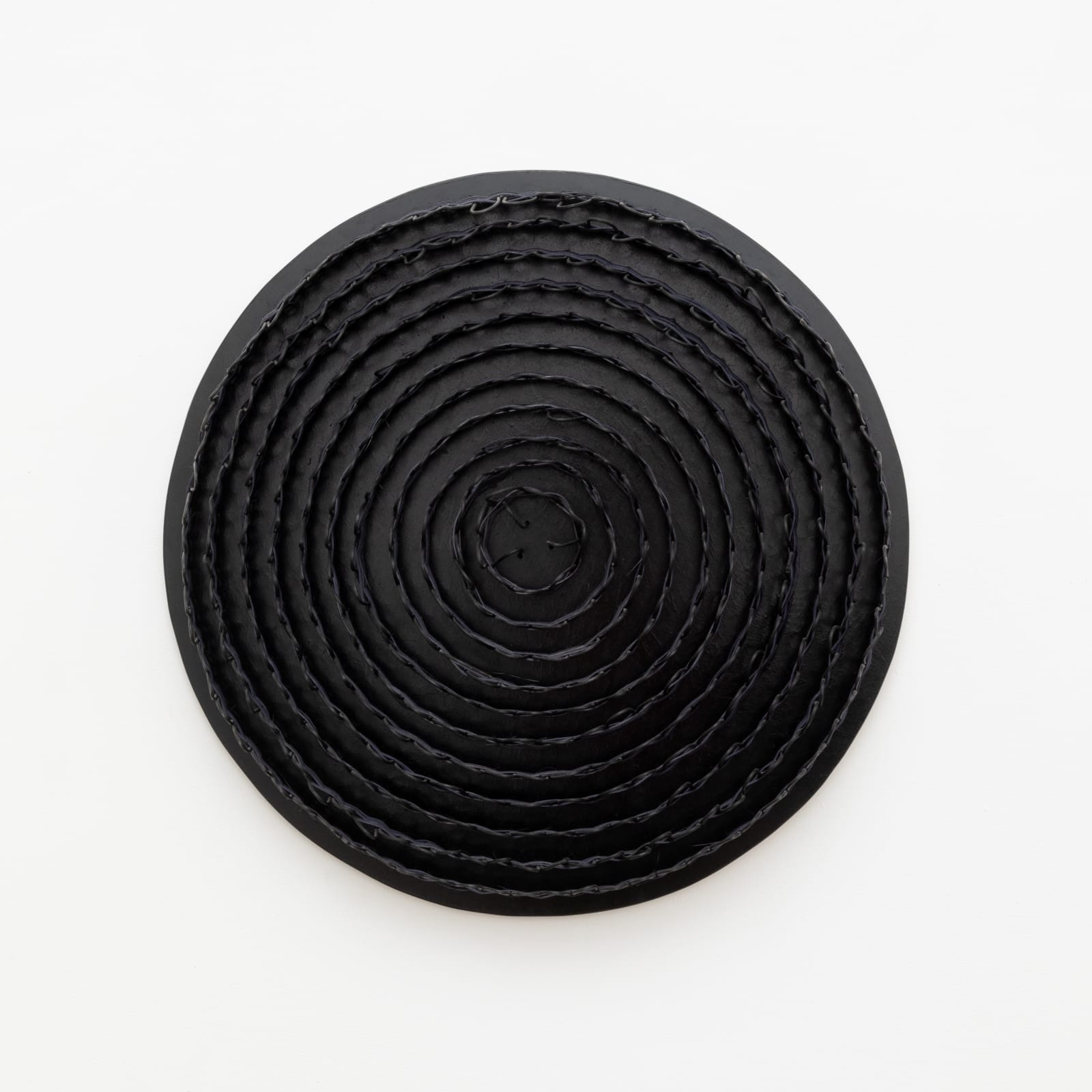 Wall-mounted sculpture consisting of a black wooden circular base with woven black reeds attached in a spiraling pattern from the center outward. 
