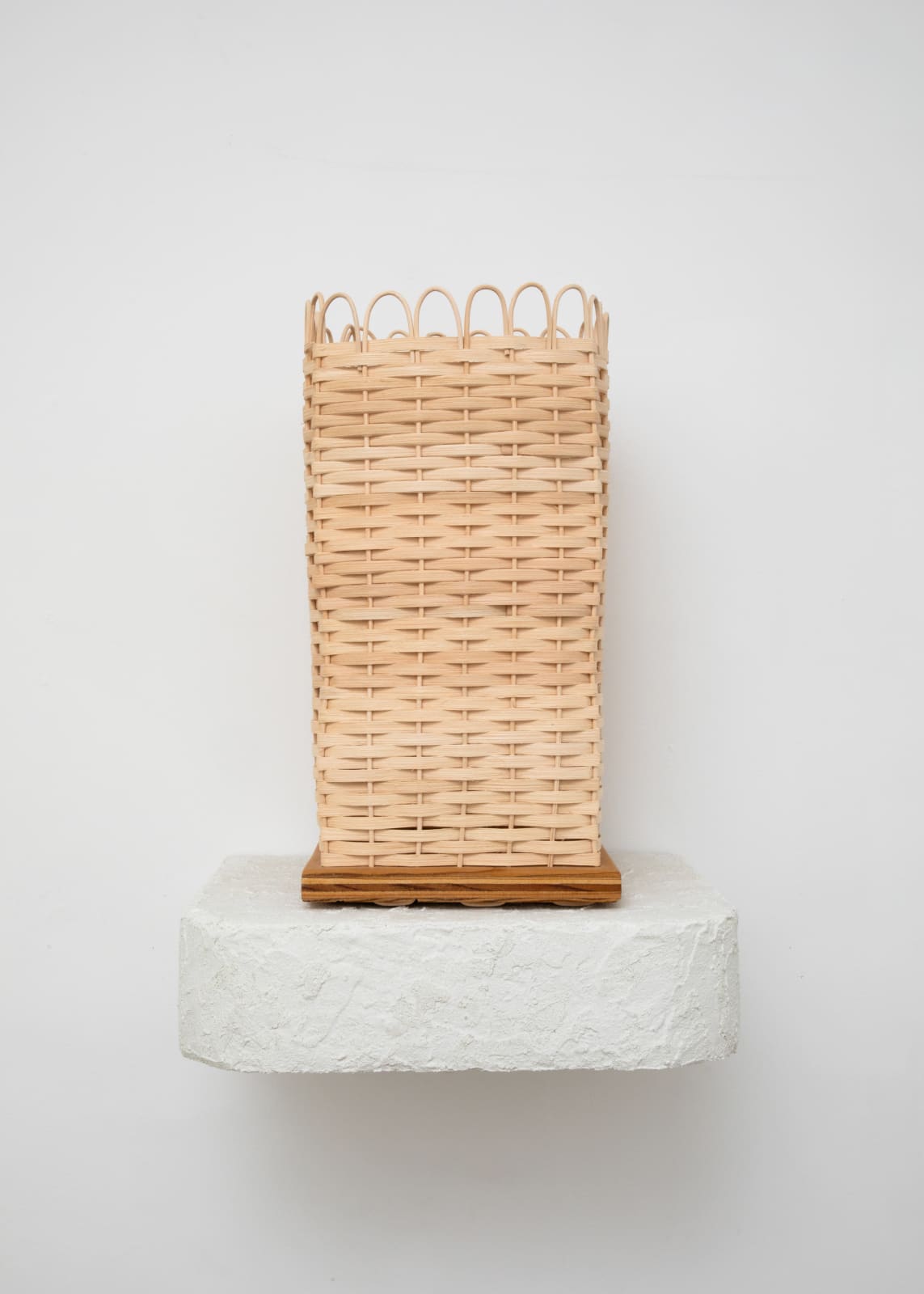 Free-standing sculpture woven with natural reed in the form of a tall square basket, that sits on top of a white stucco shelf. 