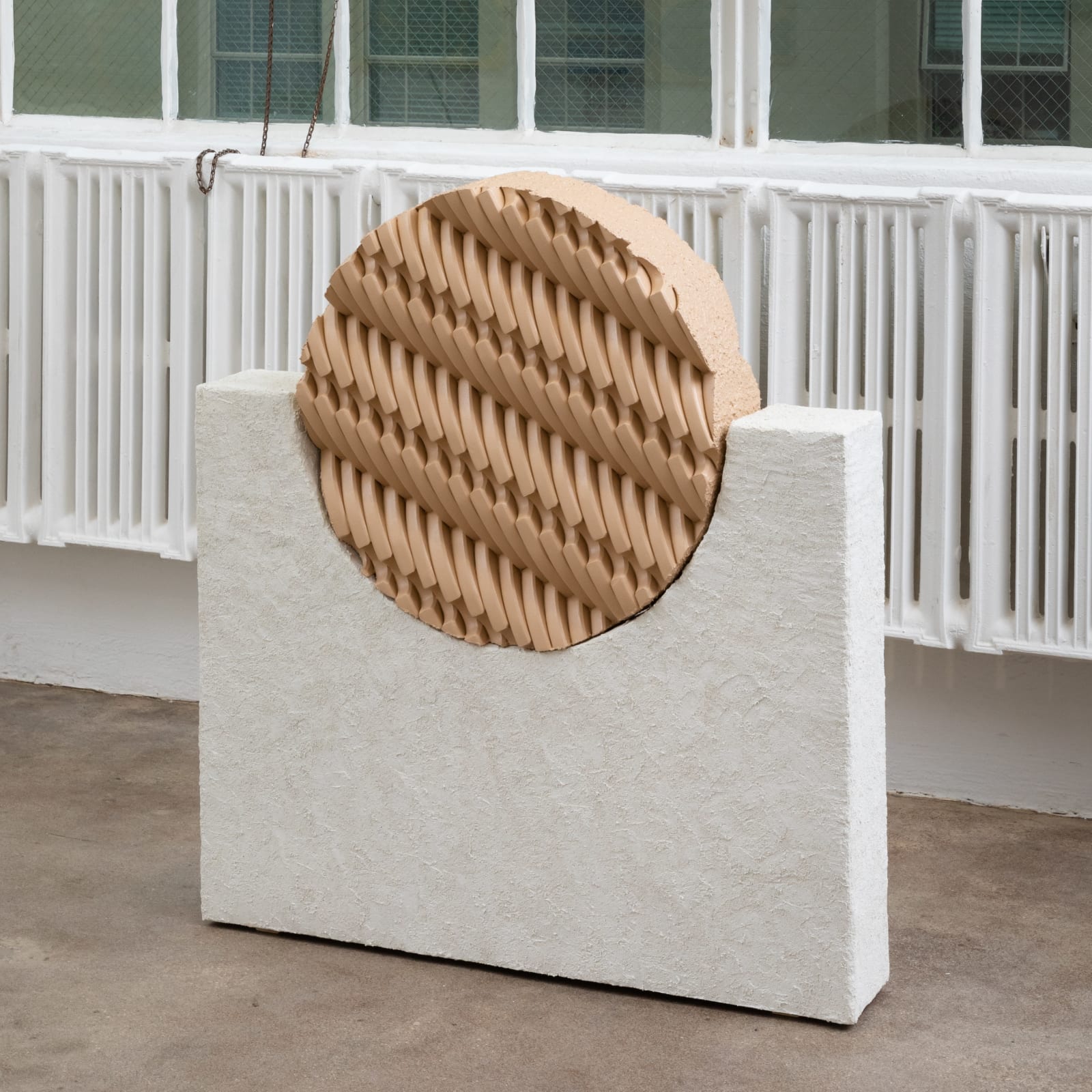 Free-standing sculpture consisting of carved polystyrene and wood pieces aggregated together to form a light tan, interlocking patterned circular form that sits in a white, tall, rectangular stucco base as a support. 