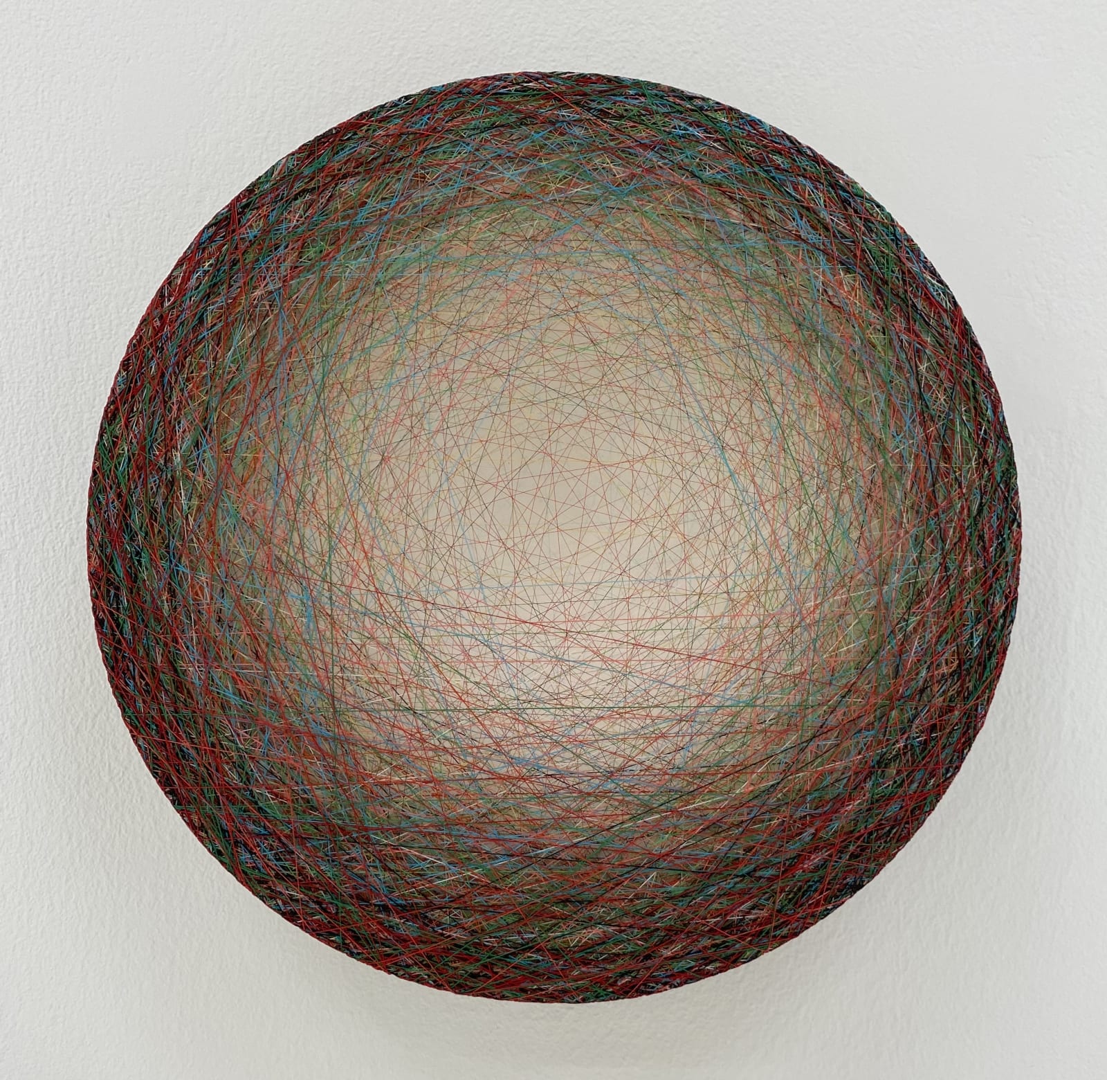 A thick concentration of layers of multi-colored threads strung across a round wooden panel create the effect of a small glowing center that is encircled by dark border