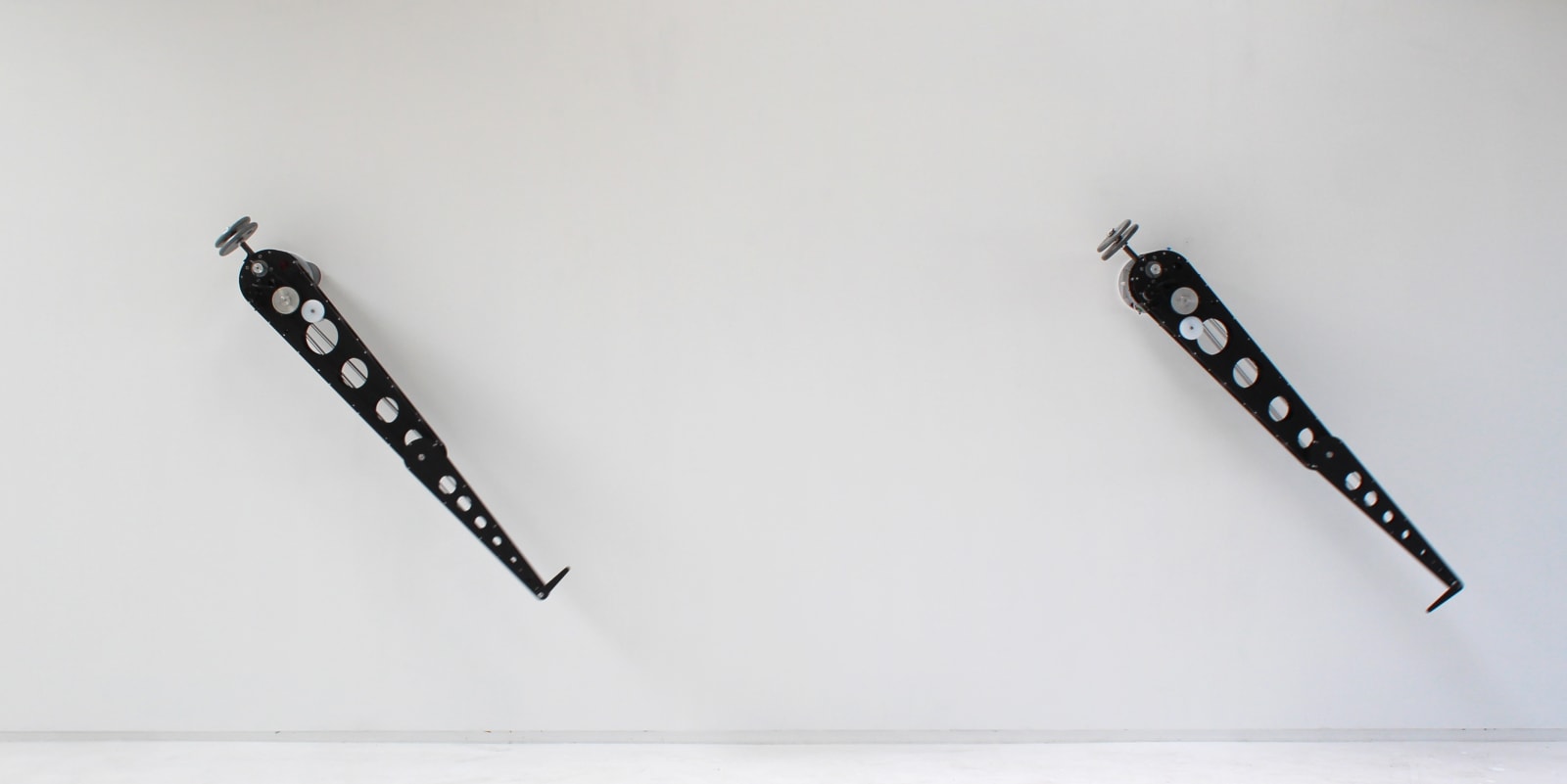 Mechanical wall mounted sculpture made out of two different black arm like structures with multiple cut holes. At the end of each arm structure a thick needle points inward towards the wall
