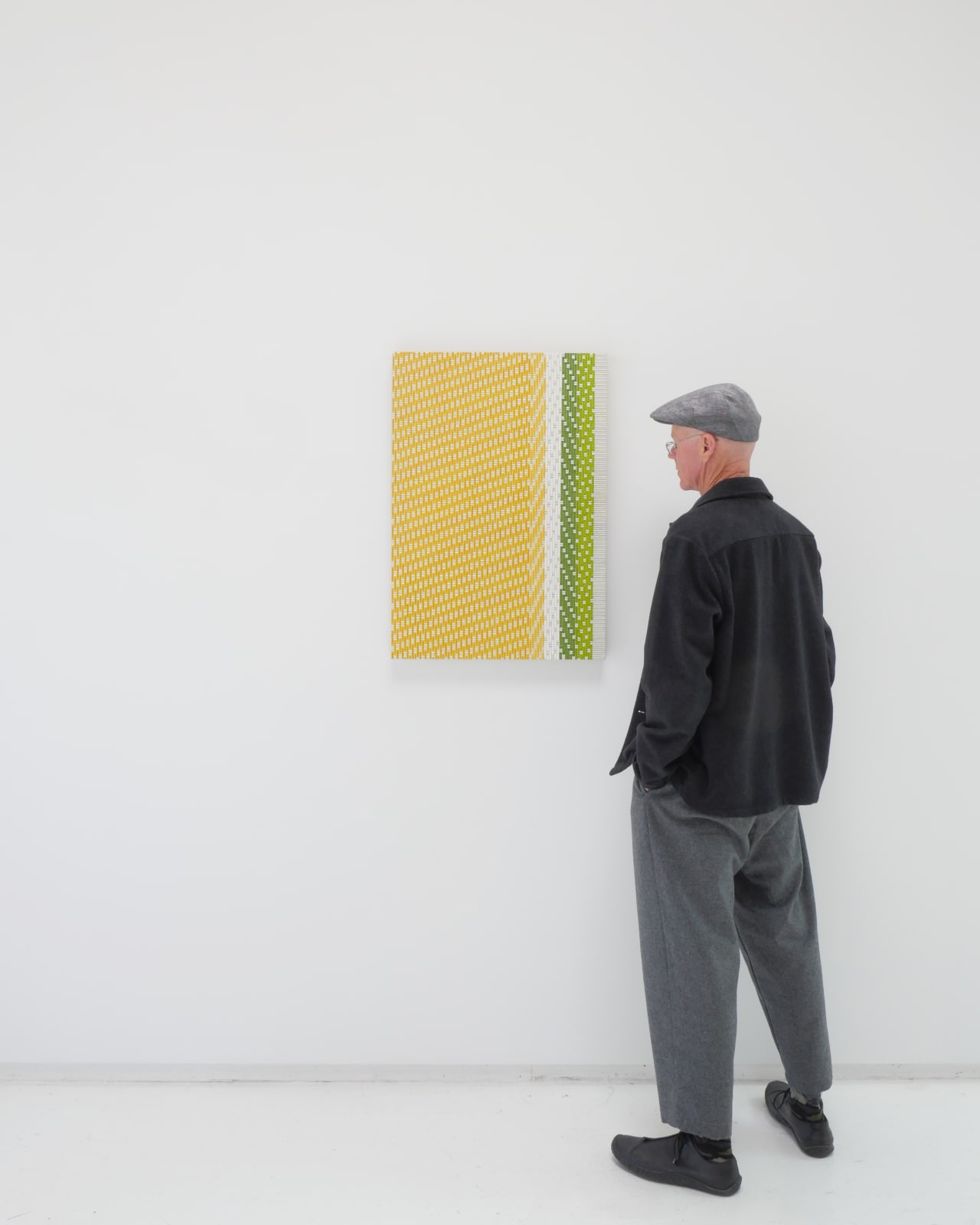 A man stands to the right and faces a yellow, white, green and grey abstract painting mimicking fabric.