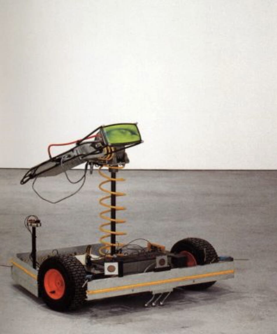 Still image of an electronic rover sculpture with two black wheels a metal boxed base custom electronics and a small elevated LCD screen. The screen displays a green colored eye