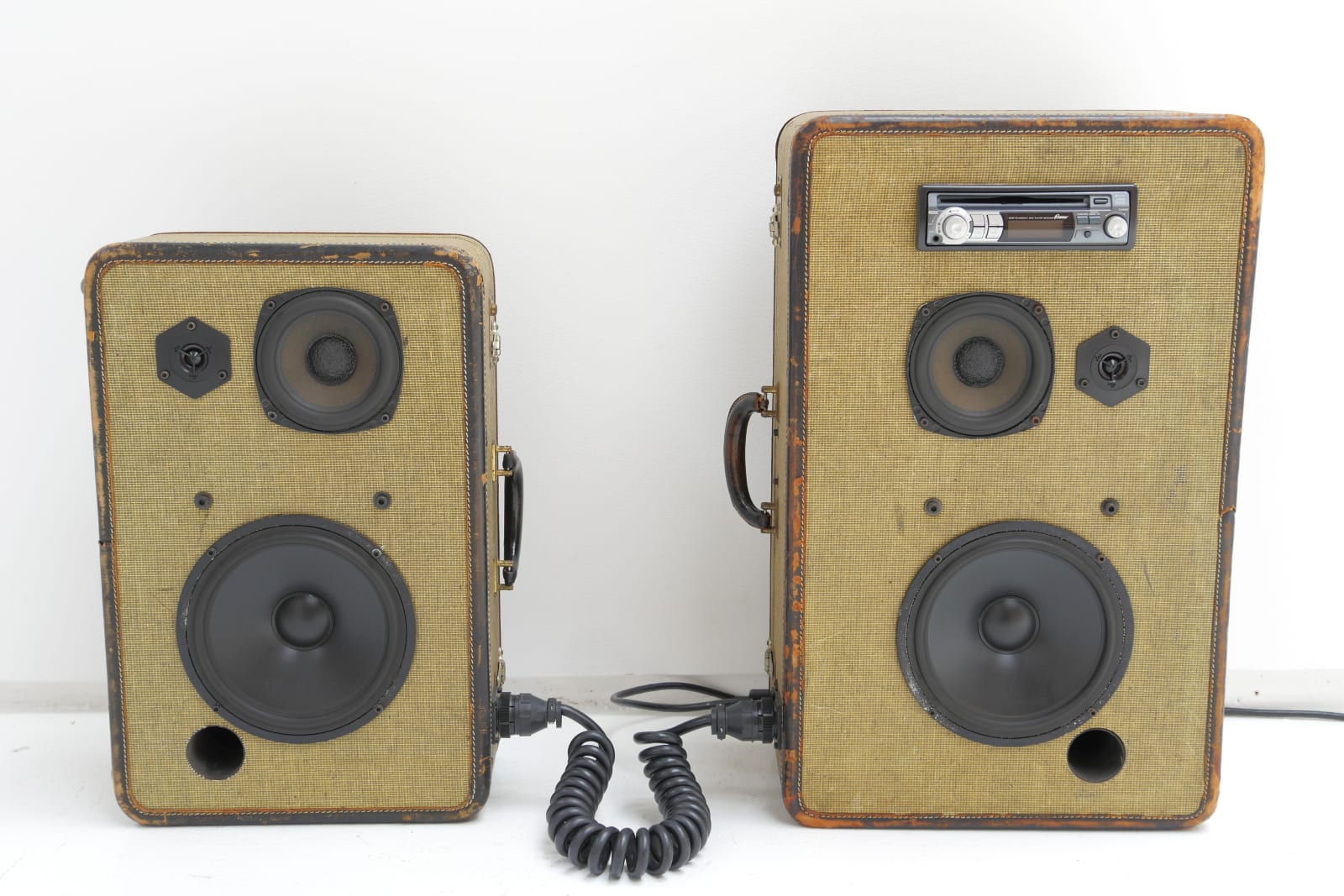 Two suitcases in two different sizes on their sides with two speakers installed on their fronts. They're connected by a coiled black cord. The larger suitcase has a stereo installed as well.