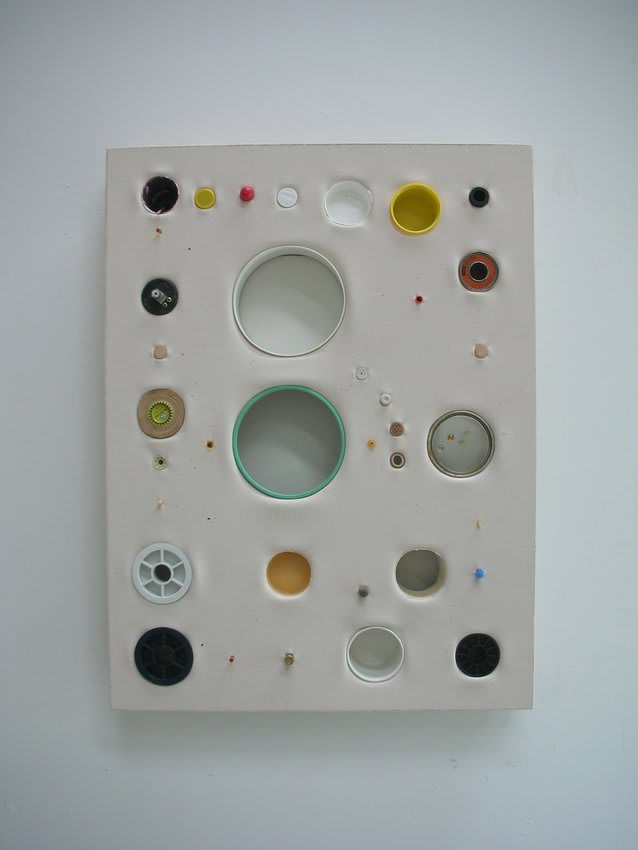 Several multi-colored circular found objects ranging in size are inserted into a pale beige painted rectangular canvas so that parts of the surface recess into depth while other portions extend beyond the canvas surface