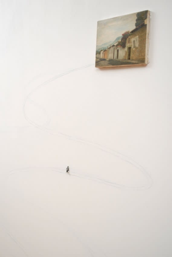 Digital photograph of an installation in which a yellowed painting of a row of rural houses is mounted onto a white wall. A string extends from the lower left corner of the painting and snakes across the surface of the wall. A tiny human figurine appears to be walking along the winding string path and up to the row of houses