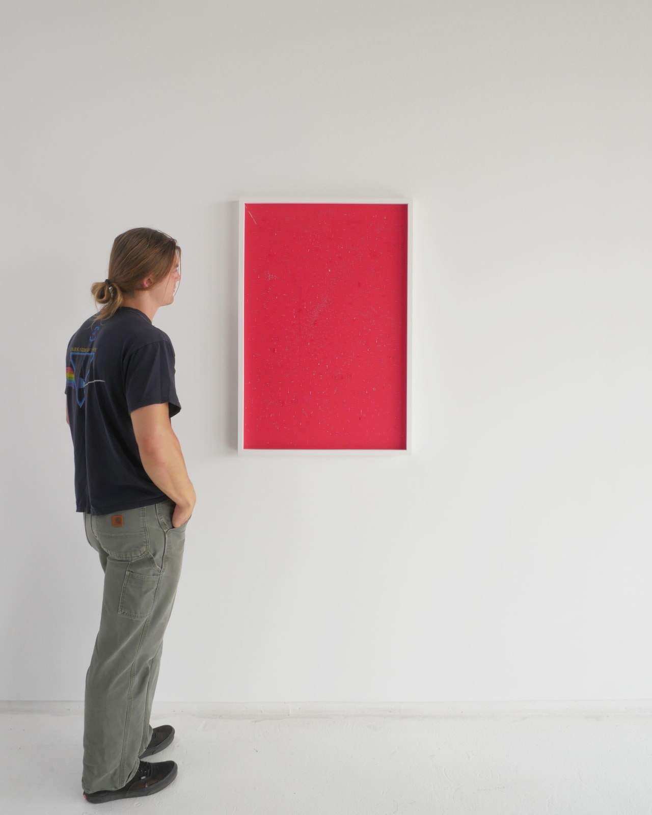 A man stands to the left and faces a red vertical rectangle in a white frame hanging on a white wall.