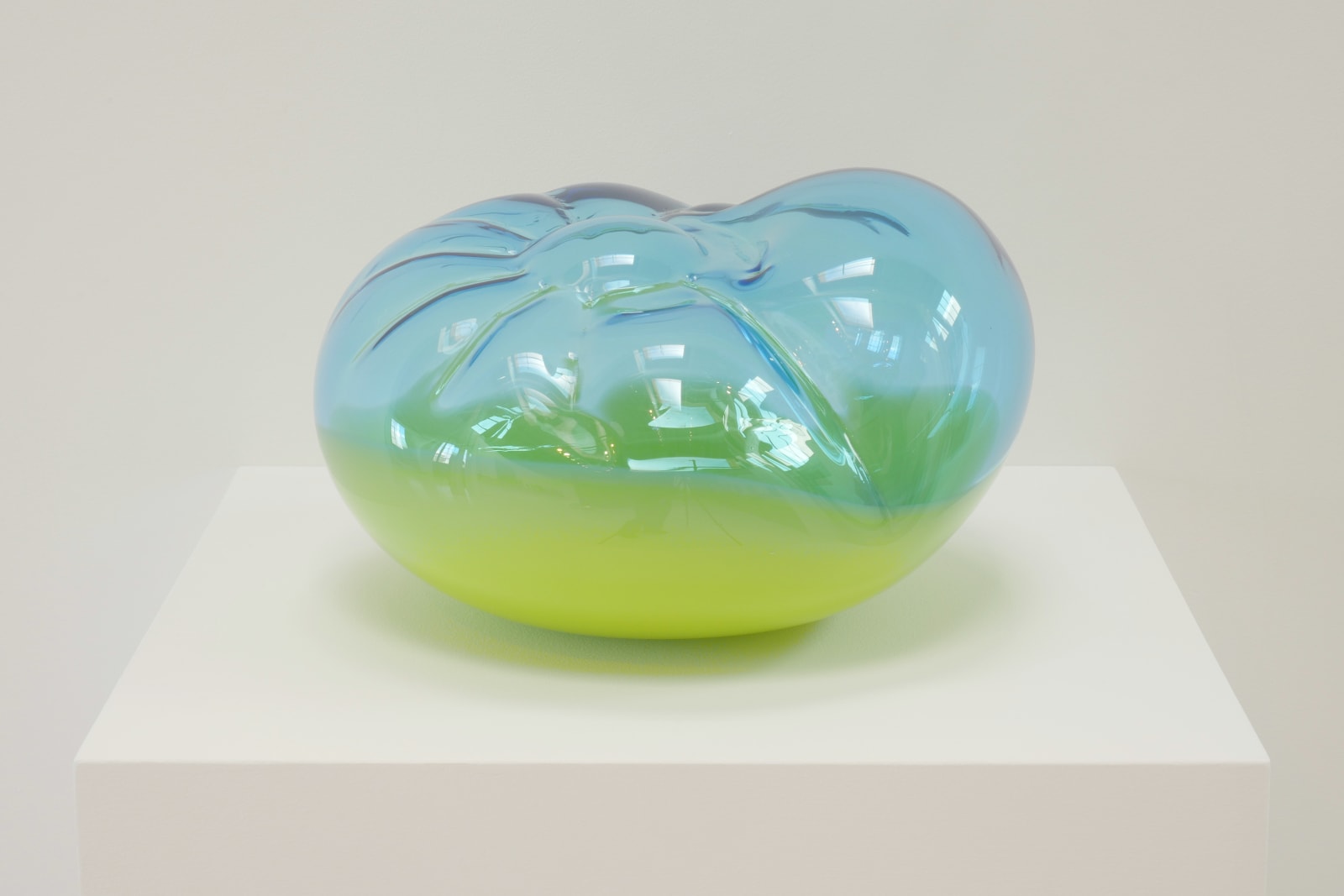 A cushion-like blown-glass sculpture, top 2/3 translucent blue, bottom 1/3 opaque yellow on a white pedestal.