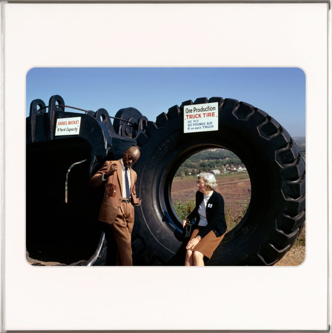 An older white woman sits on the edge of a large tire while artist, Omar Diop chats with her. By Lee Shulman & The Anonymous Project.