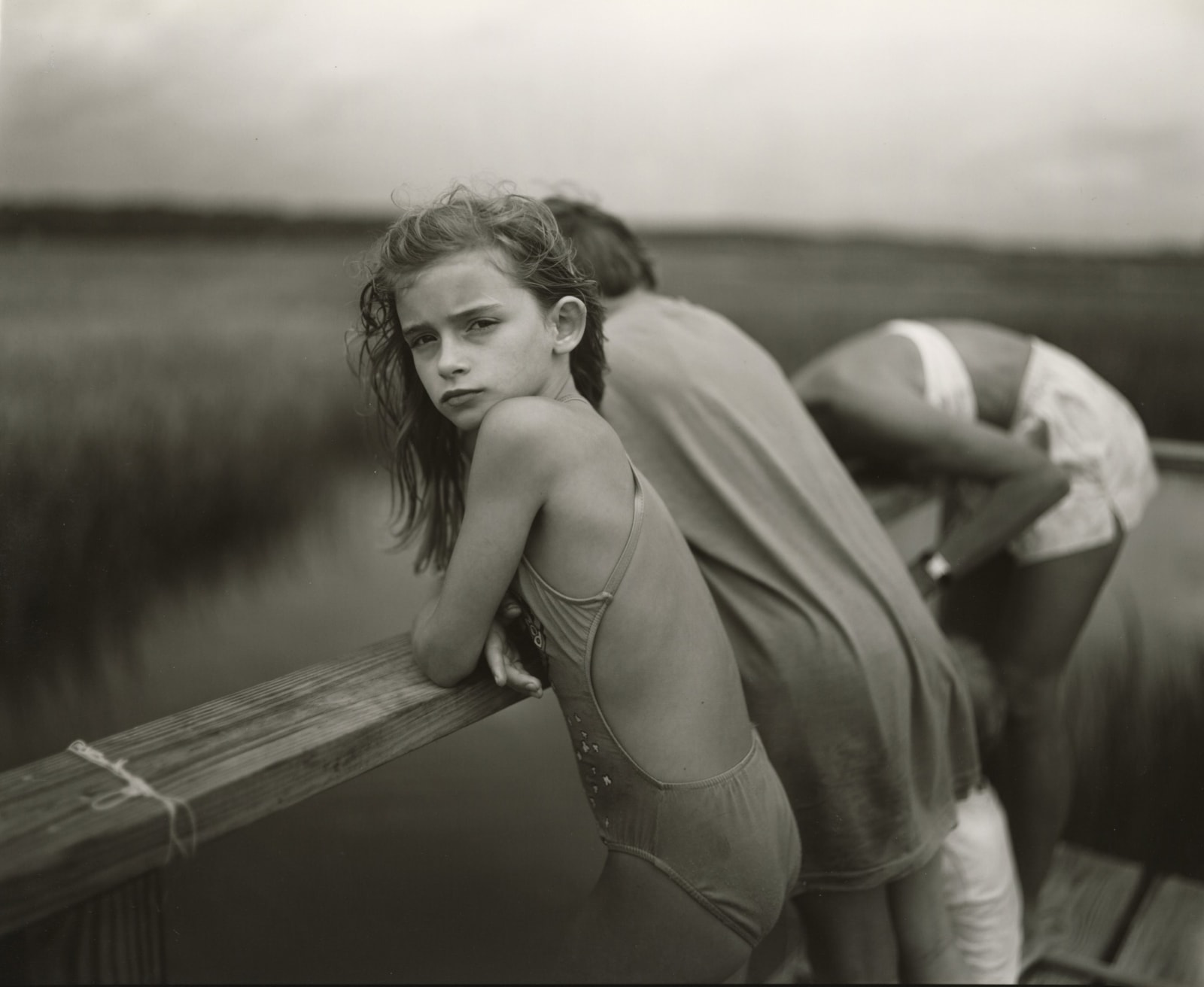 young girl in bathing suit leaning over lake fence, hair blowing in wind, by Sally Mann from the Immediate Family series