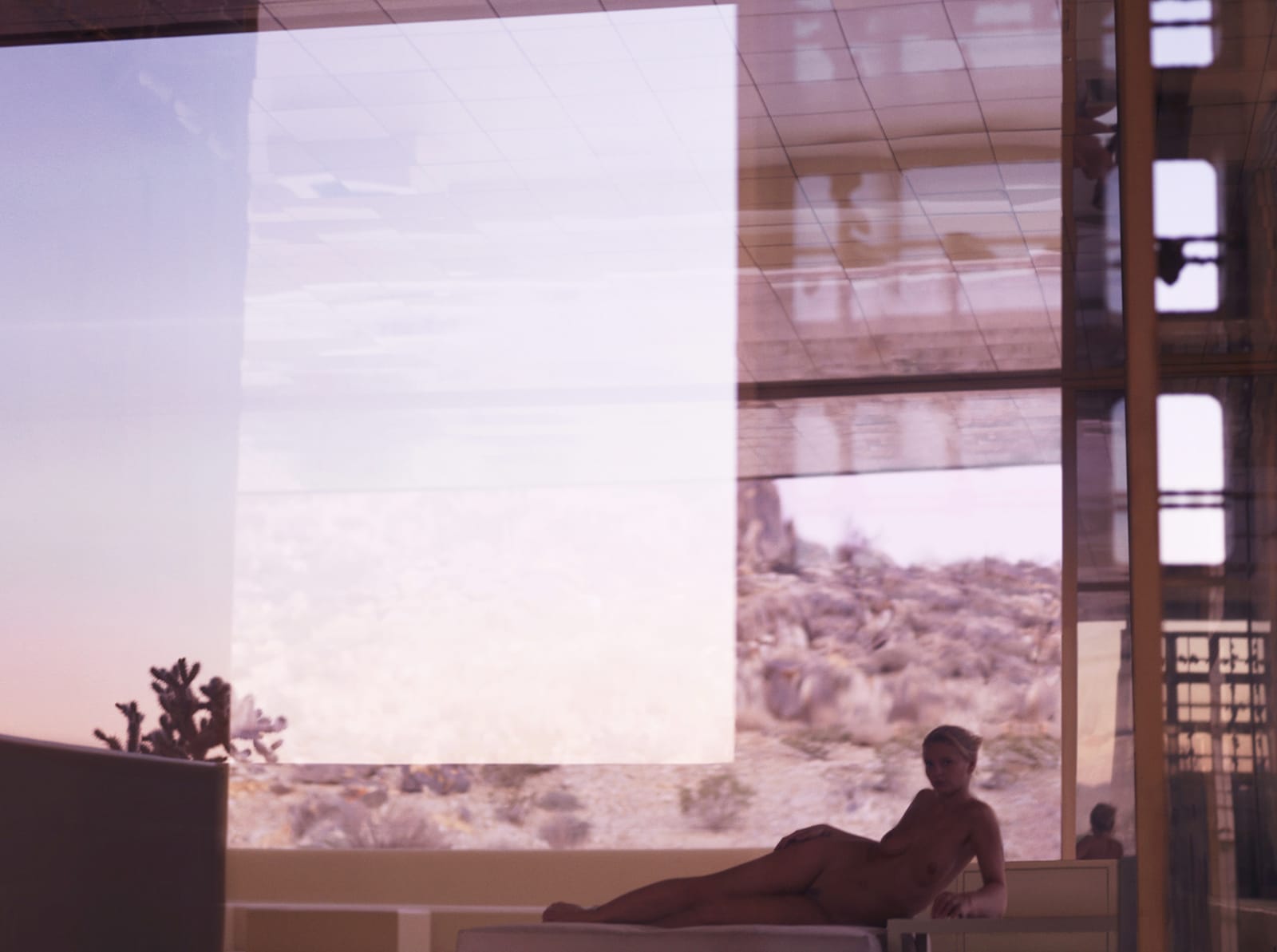 woman reclining in glass modernist house with reflections and California desert in rosy hue behind, by Mona Kuhn
