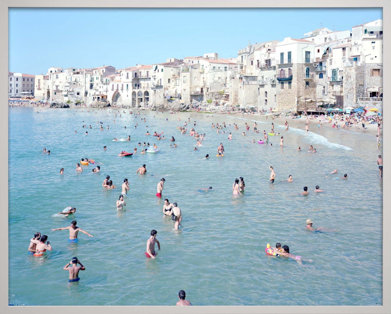 Beachgoers wade and swim in clear blue water near a historic seaside town; photograph by Massimo Vitali.