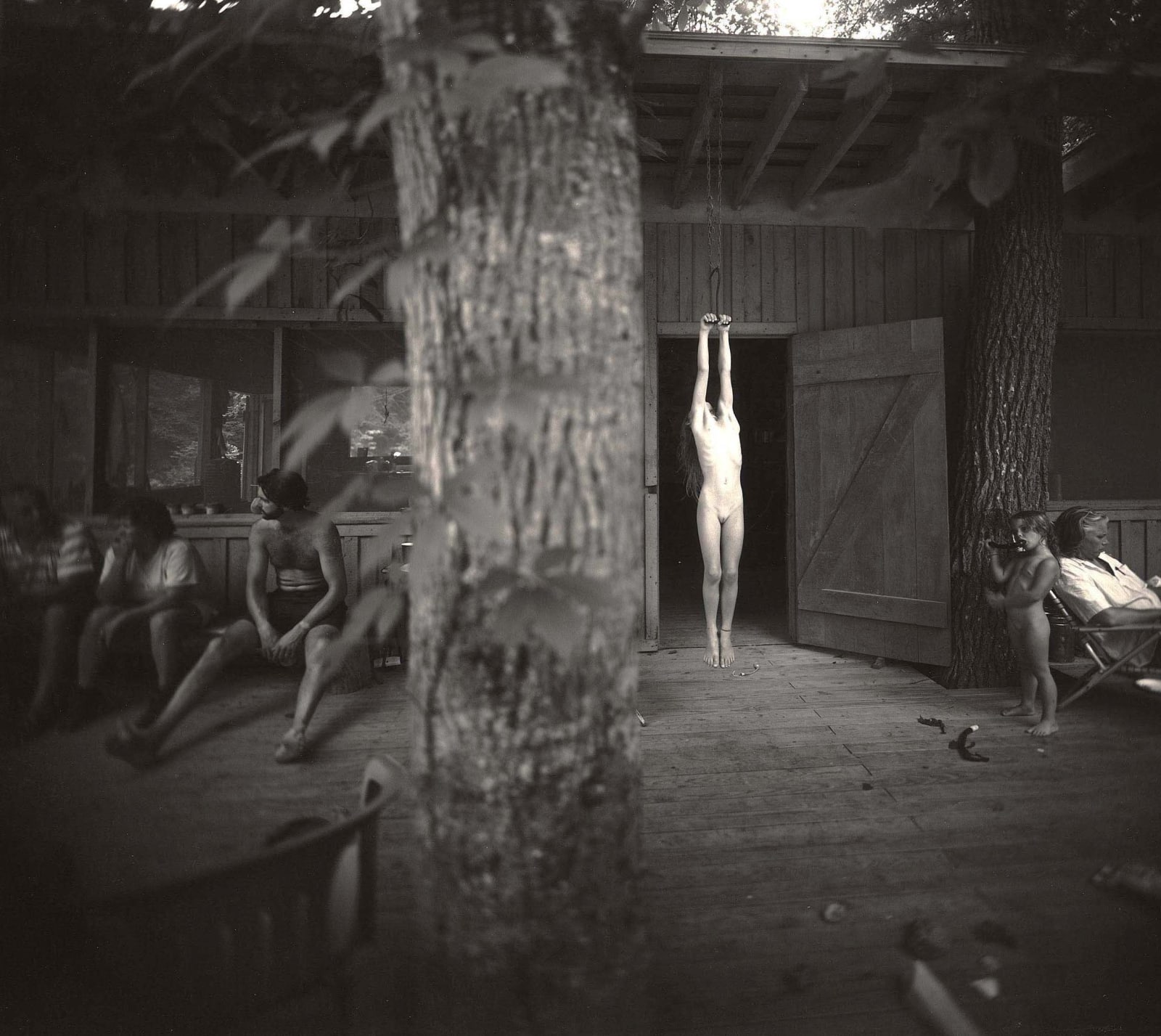 Jessie Mann nude hanging on a hayhook with Larry Mann and Virginia Mann and tree in foreground, from the Immediate Family series by Sally Mann