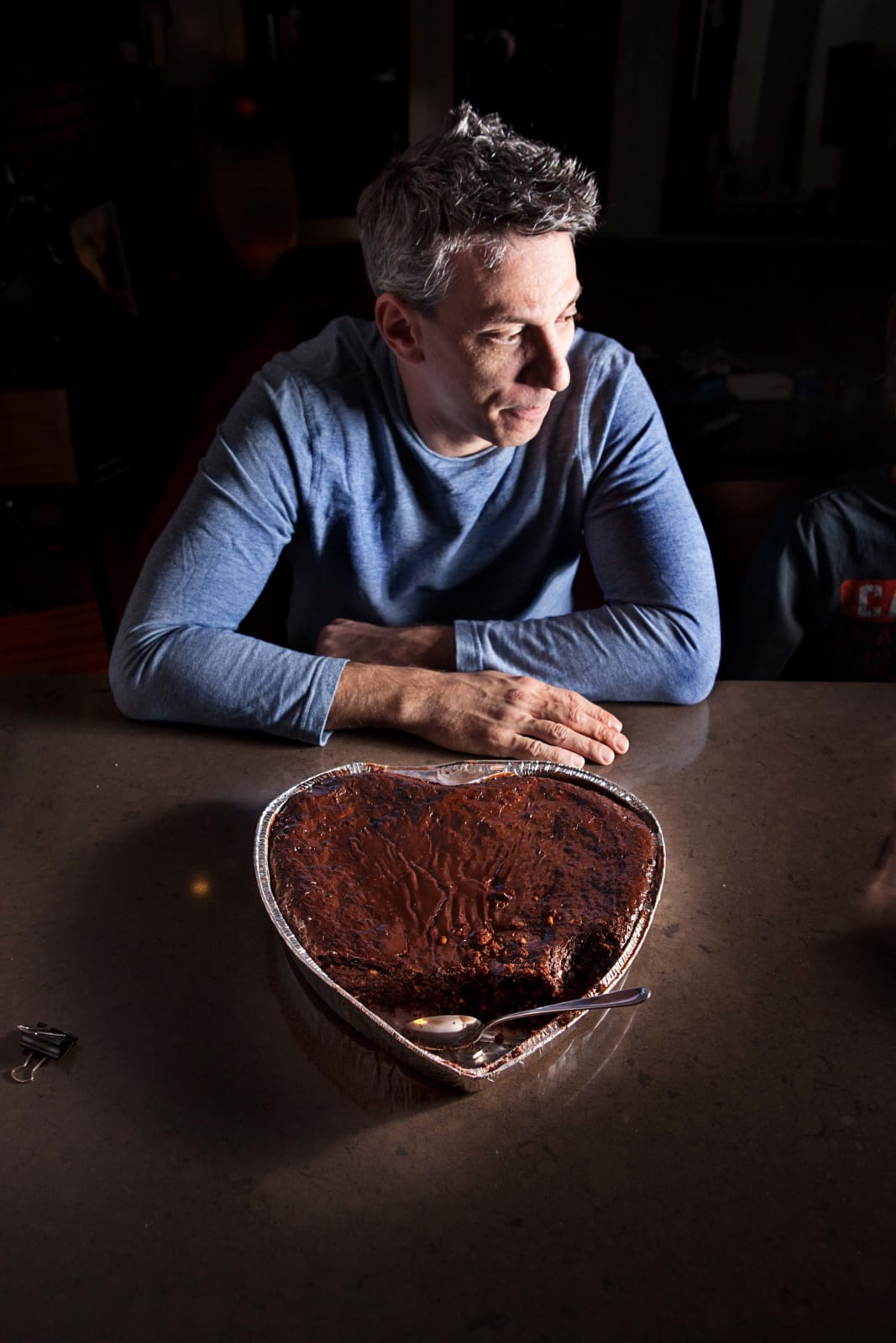 Elinor Carucci photograph of the artist's husband sitting at the table in a blue sweater with a heart shaped chocolate cake