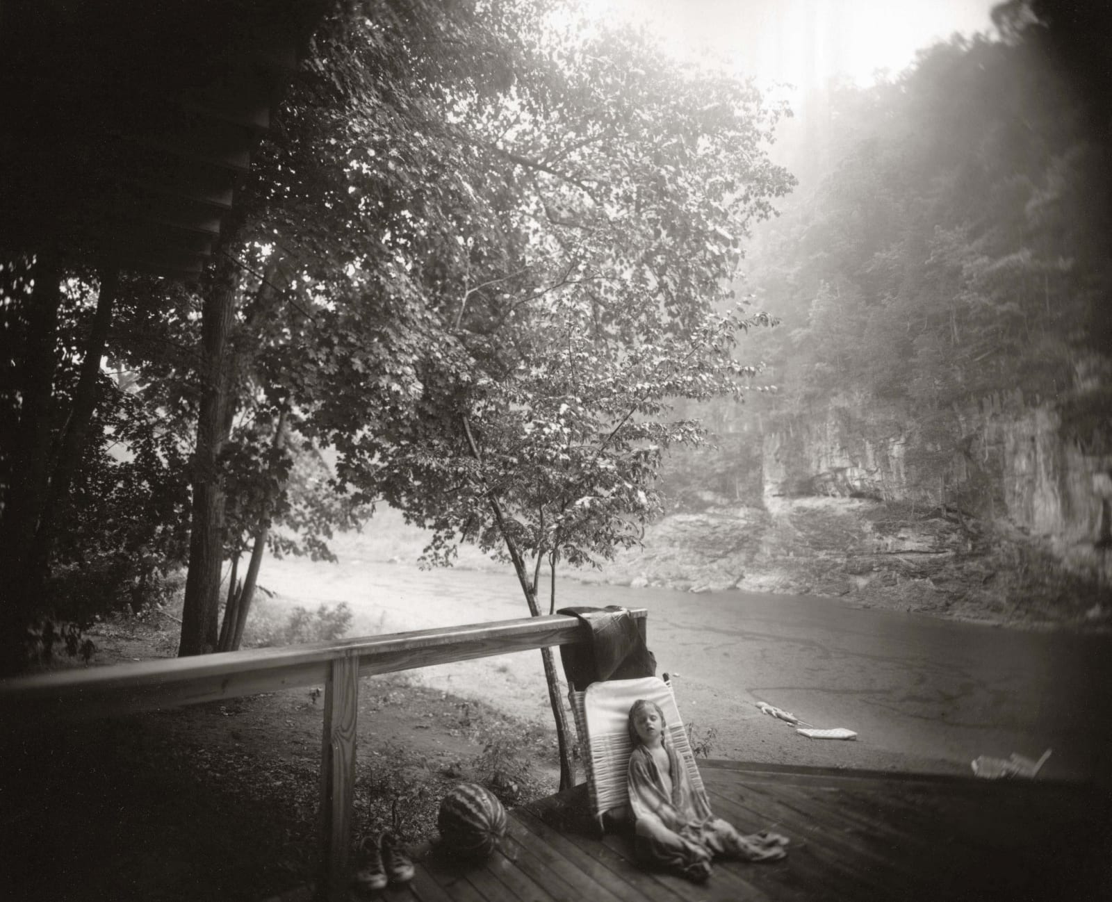Virginia sleeping on patio with alligator swim toy on river's edge in background, from the Immediate Family series by Sally Mann