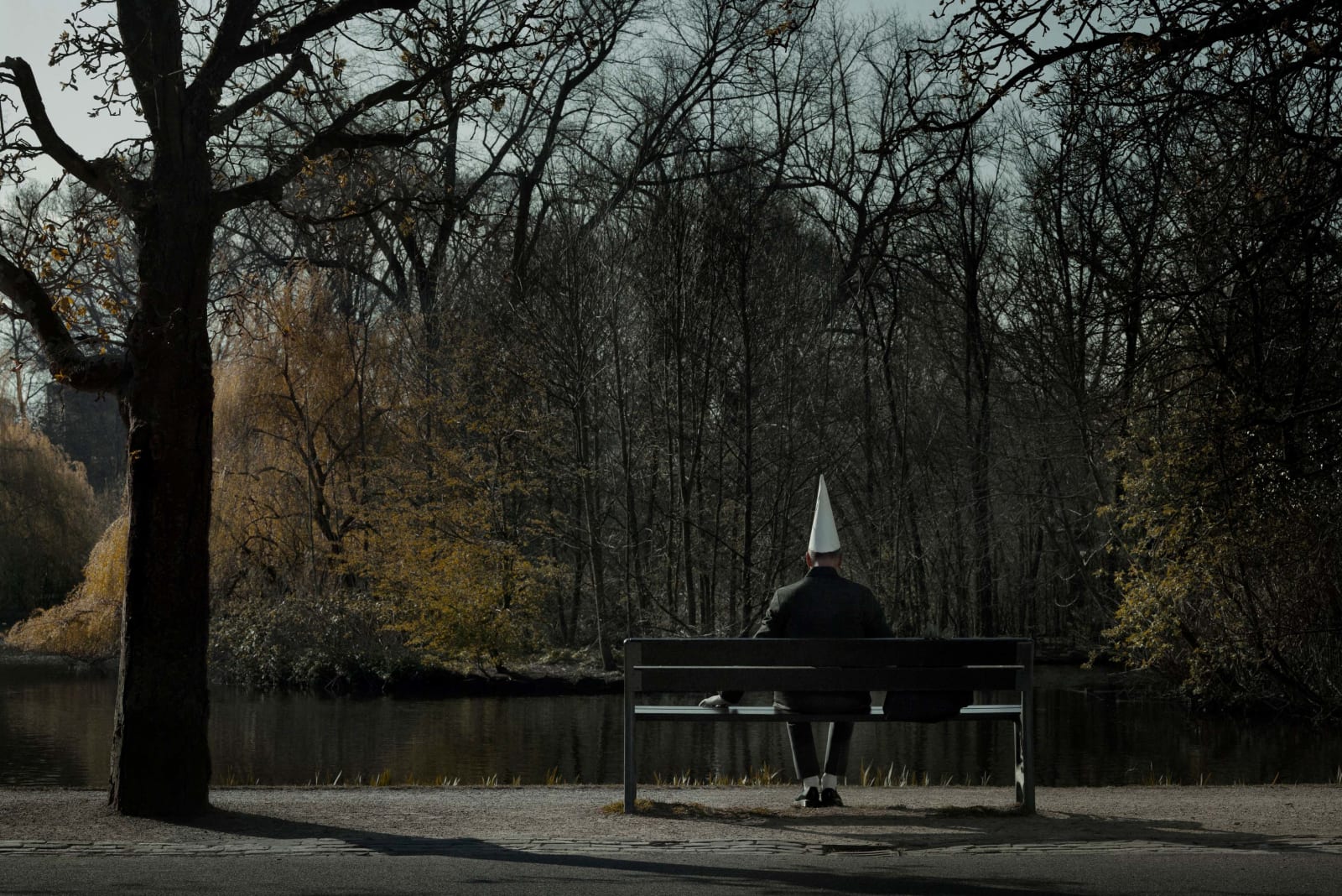 Erwin Olaf in dunce cape sitting on park bench facing water and autumnal foliage, by Erwin Olaf