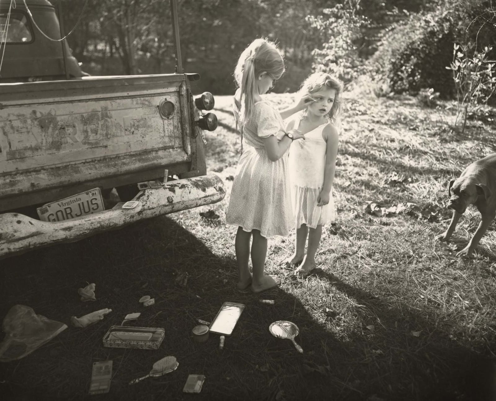 Jessie Mann helping Virginia Mann with her make-up as young girls, mirrors on the ground and truck with Gorjus license plate next to the two girls, by Sally Mann