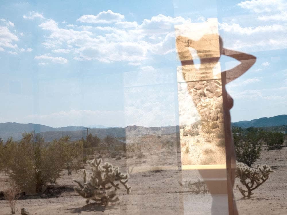 Mona Kuhn photograph of nude woman in desert behind glass, with refracted light and succulents reflected on top of her silhouette, clouds in blue sky