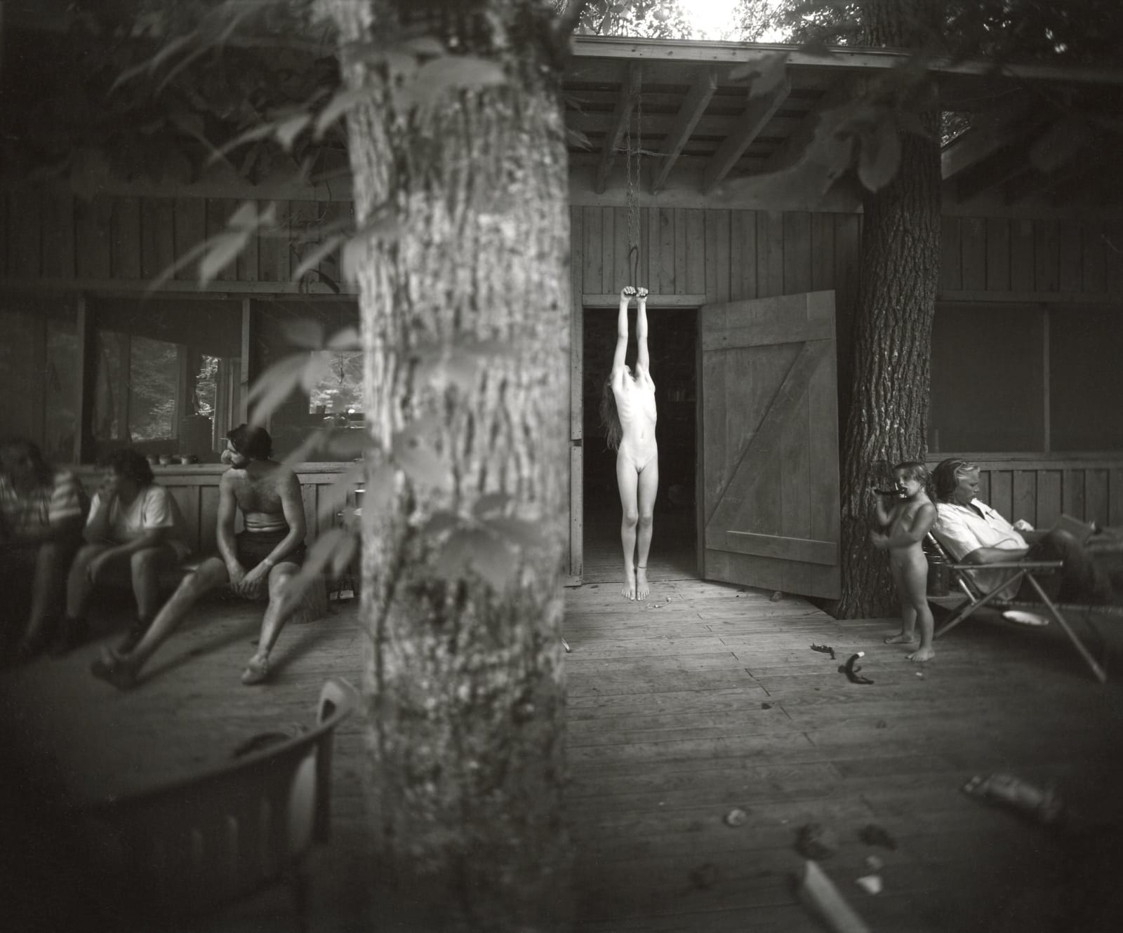 Jessie Mann nude hanging on a hayhook with Larry Mann and Virginia Mann and tree in foreground, from the Immediate Family series by Sally Mann