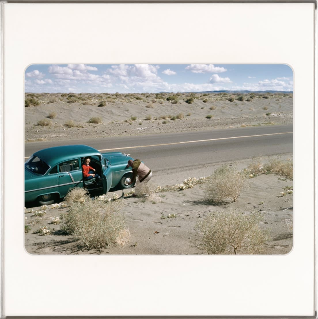 A white woman sits in the passenger seat of a turquoise car while artist, Omar Diop bends down to check the front tire. By Lee Shulman & The Anonymous Project.