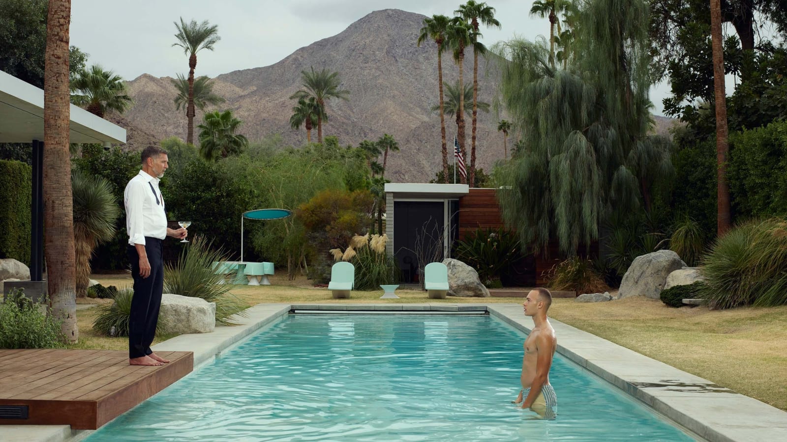 Young man standing in pool in Palm Springs, looking at older man holding martini glass at edge of pool, by Erwin Olaf