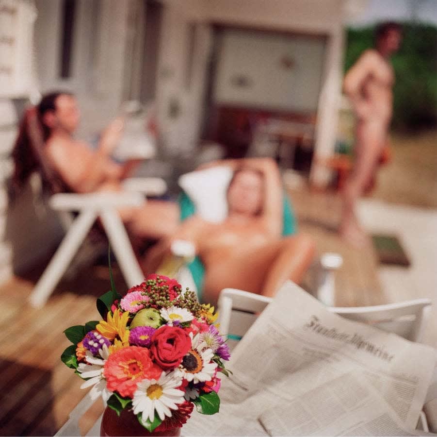 a bouquet of flowers and newspaper in the foreground with three nude people sunbathing out of focus, by Mona Kuhn