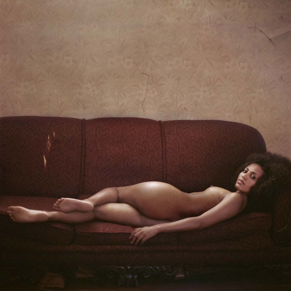 Mona Kuhn photograph of nude woman reclining on burgundy couch looking at viewer