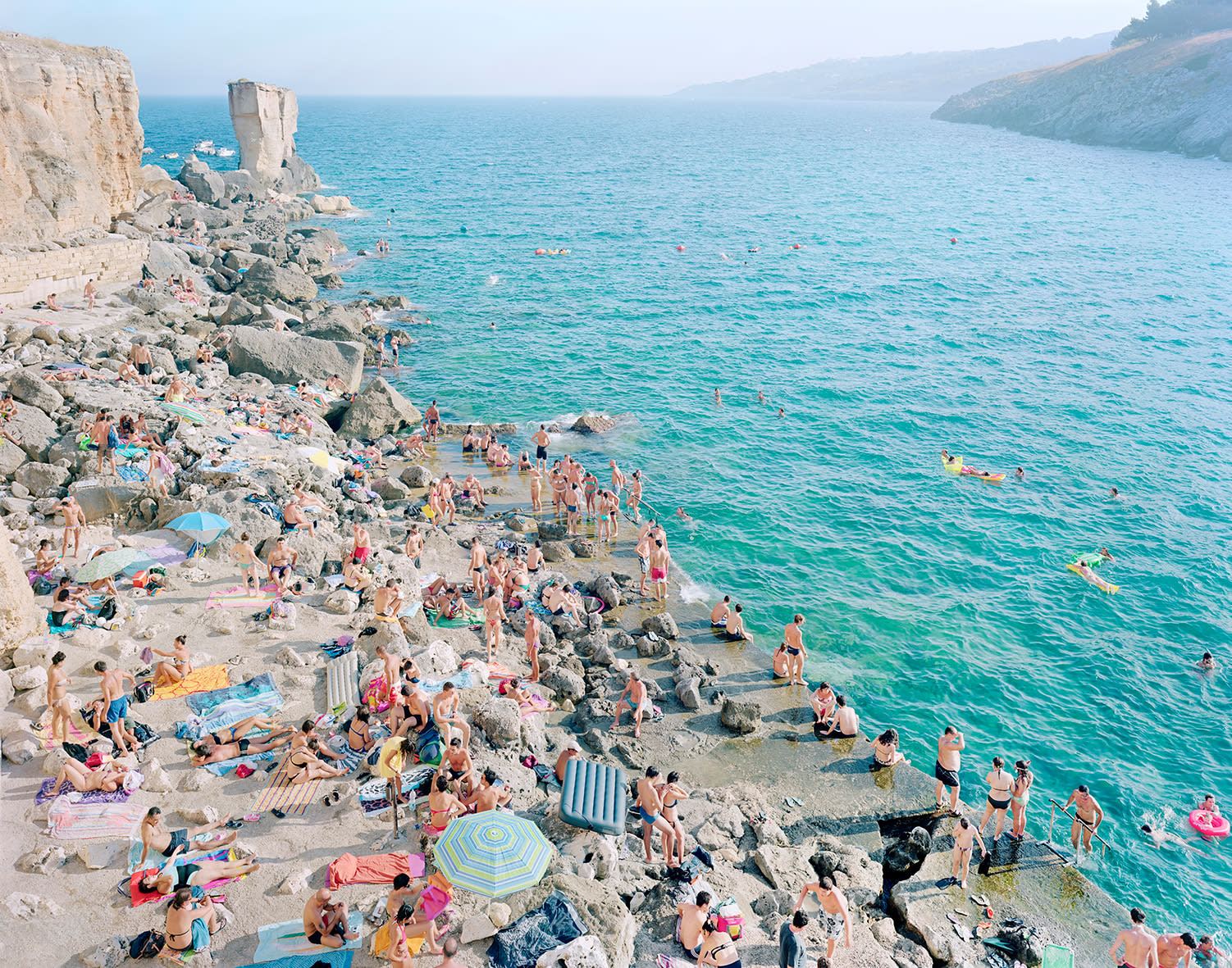 Crowds relax on rocky shores and swim in turquoise water under bright sun; photograph by Massimo Vitali.