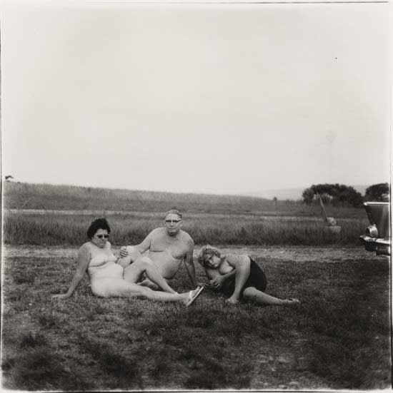 Diane Arbus photograph of a family of nudists, three nude people sitting in grass with car fin barely in view