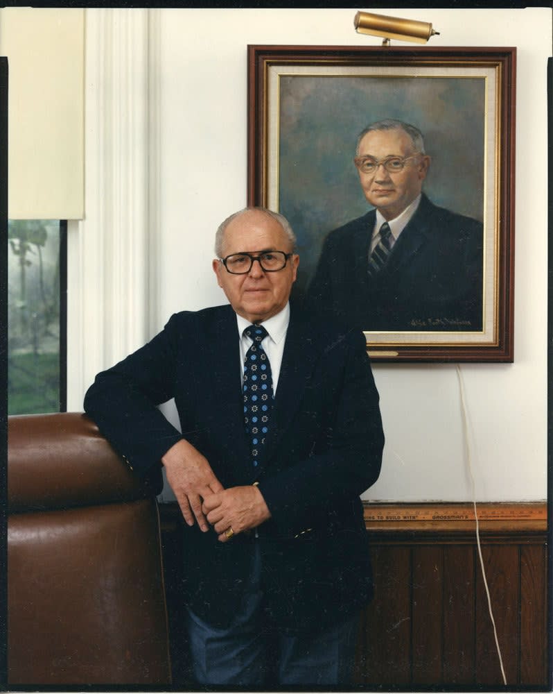 Bruce Wrighton, Man in dark suit in front of framed portrait on wall