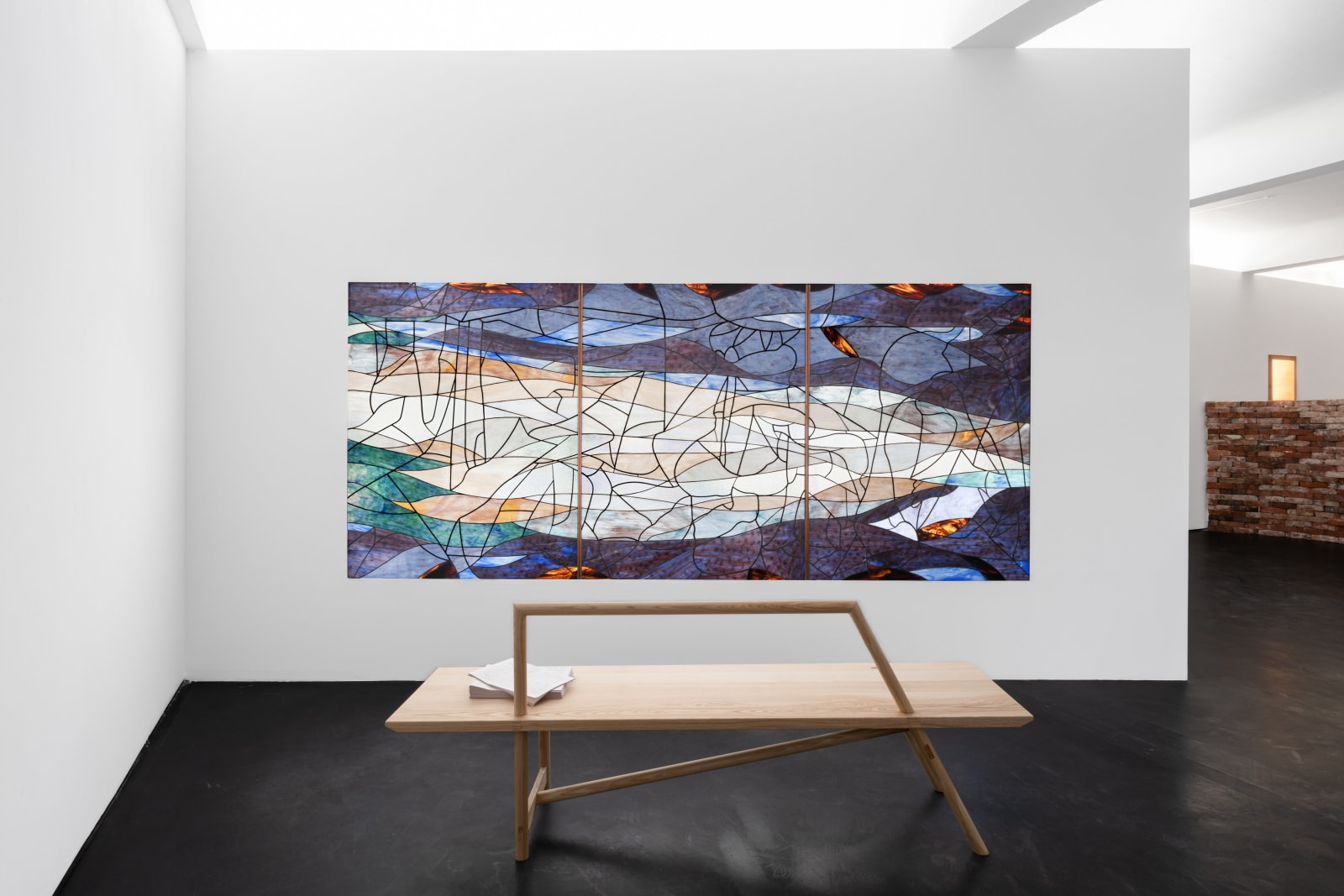 Room with a dark blue, green and orange stained glass window made up of 3 panels against a plain wall, a brick wall behind it and a wooden bench in front of it with a stack of papers on it. Rindon Johnson Clattering, 2021 Stained glass work (3 panels) and ash wood bench Glass work: 360 x 158 cm 141 3/4 x 62 1/4 inches Bench: 200 x 49 x 80 cm 78 3/4 x 19 1/4 x 31 1/2 inches