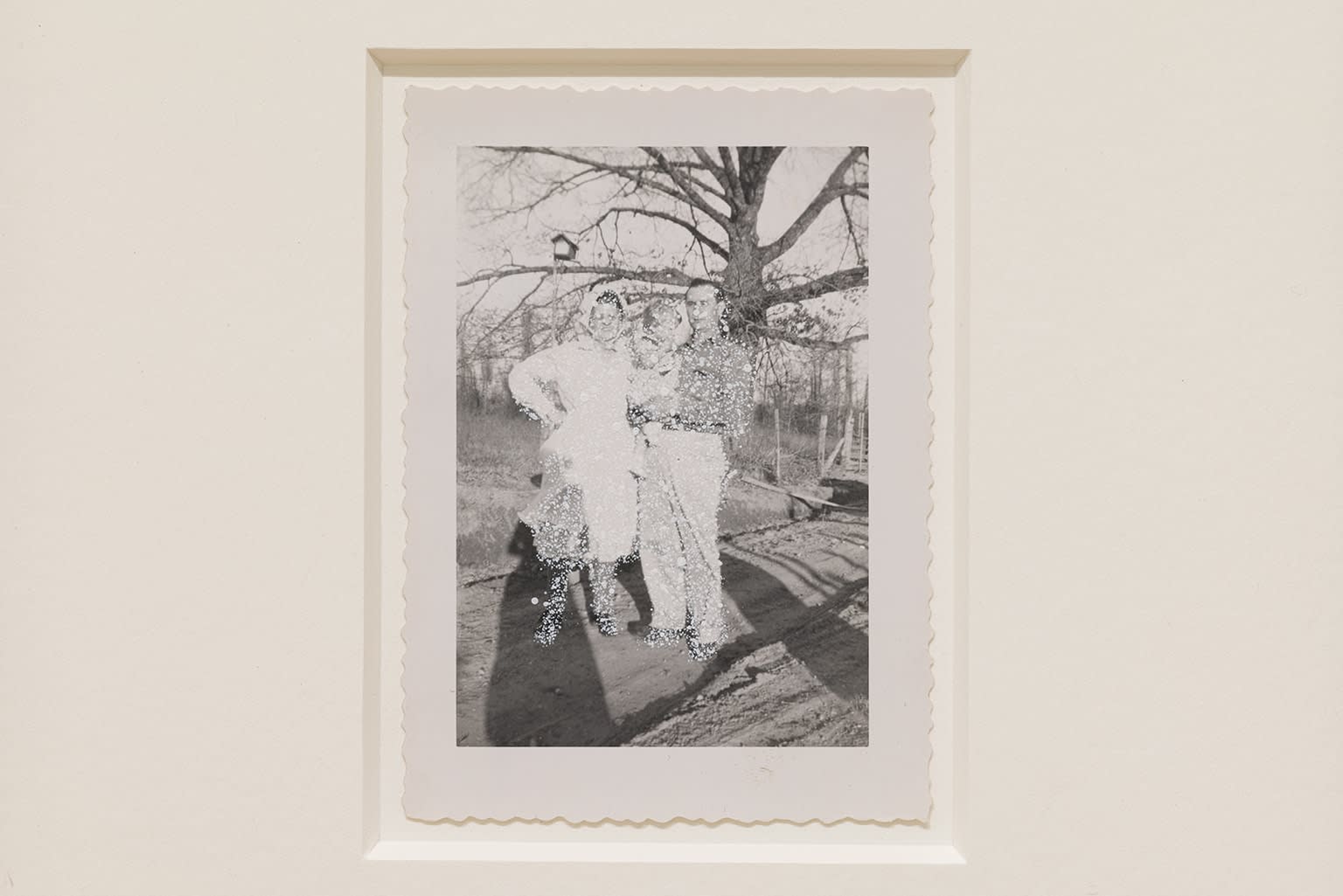 John Latour, Woman with bonnet posing with her son holding his daughter. Bird feeder hanging from a nearby branch, 2025