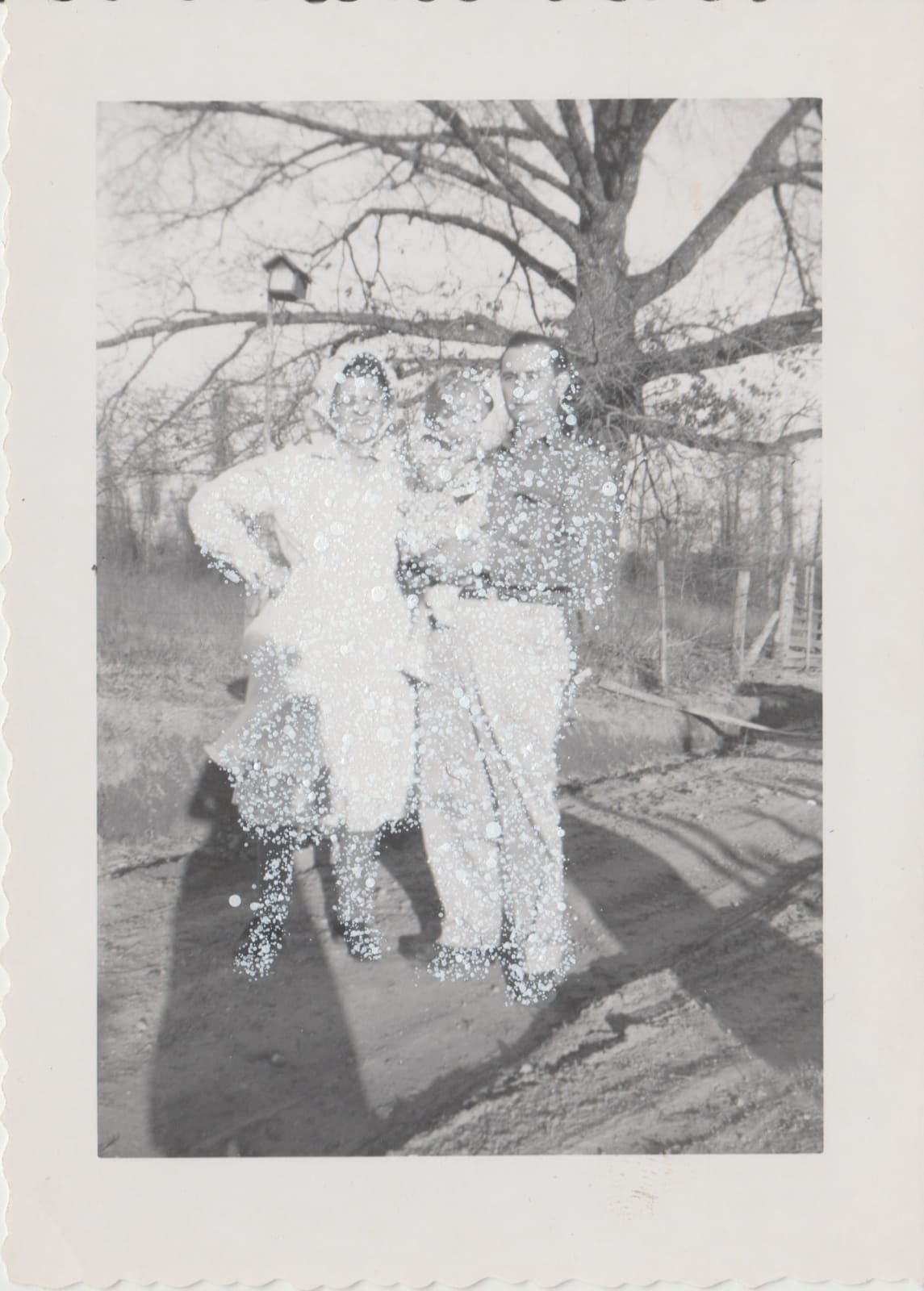 John Latour, Woman with bonnet posing with her son holding his daughter. Bird feeder hanging from a nearby branch, 2025
