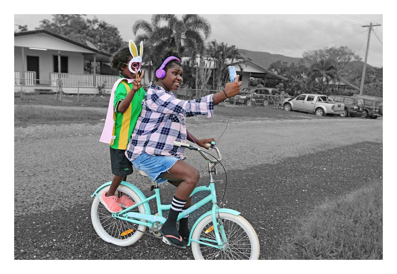 A girl on a bike taking a selfie on her phone of her and her little brother who is standing on the back of the bike. Aboriginal artwork photography which showcases indigenous people in a natural light. By Naomi Hobson, Aboriginal-Australian female artist.