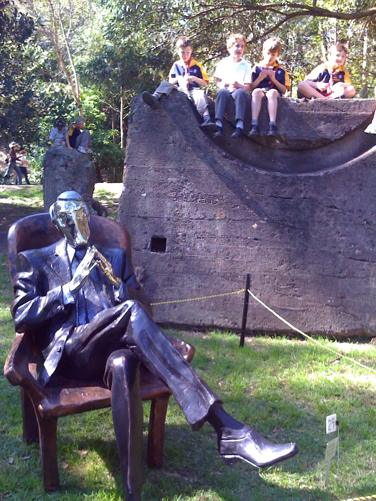 A resin sculpture titled Chairman depicting a seated male figure in a suit. The man is portrayed in a thoughtful pose, leaning back slightly in a high-backed chair with his legs crossed and hands clasped together. The figure’s head and hands have chrome finishes, contrasting with the darker patina of the suit and chair. The piece exudes authority and contemplation. The sculpture is displayed in roped off area in a park next to a large sandstone boulder which four children are sitting on with their legs dangling over the side.