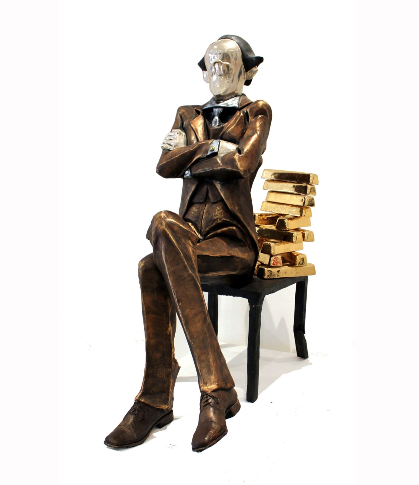 A large sculpture of a banker seated on a chair with a backrest made of stacked gold bullion. The figure, dressed in a formal suit with arms crossed, has a polished, reflective face that adds a surreal element to the piece. One chair leg is visibly bending under the weight of the gold, symbolizing the burden of wealth.
