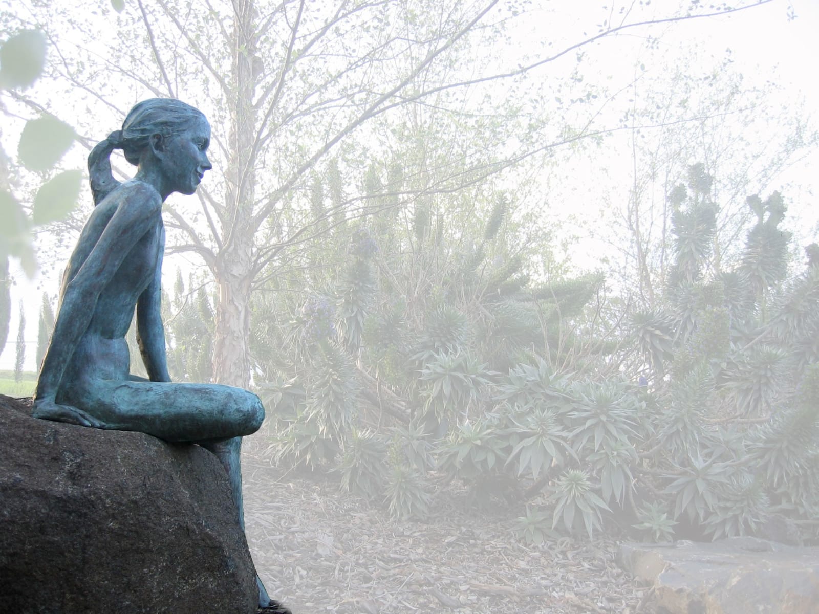 A bronze statue of a girl sitting on a large boulder, set in an outdoor garden. She gazes contemplatively at her surroundings, with her hair tied back, blending naturally with the misty greenery and shrubs in the background.