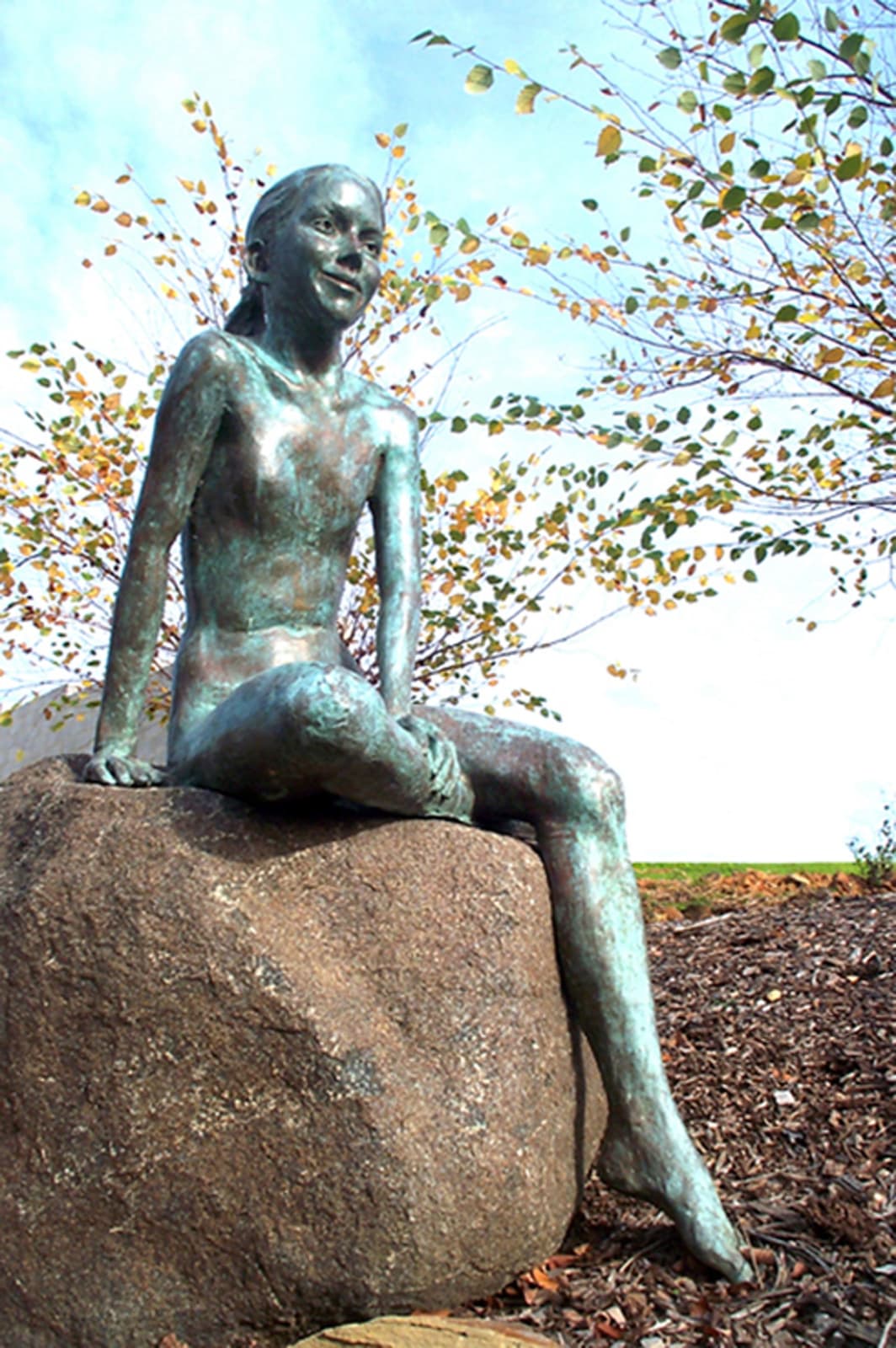 A life-size bronze sculpture of a young girl sitting on a smooth boulder outdoors, surrounded by golden autumn leaves. The girl’s playful expression and relaxed posture exude a sense of peace and harmony with the natural environment.