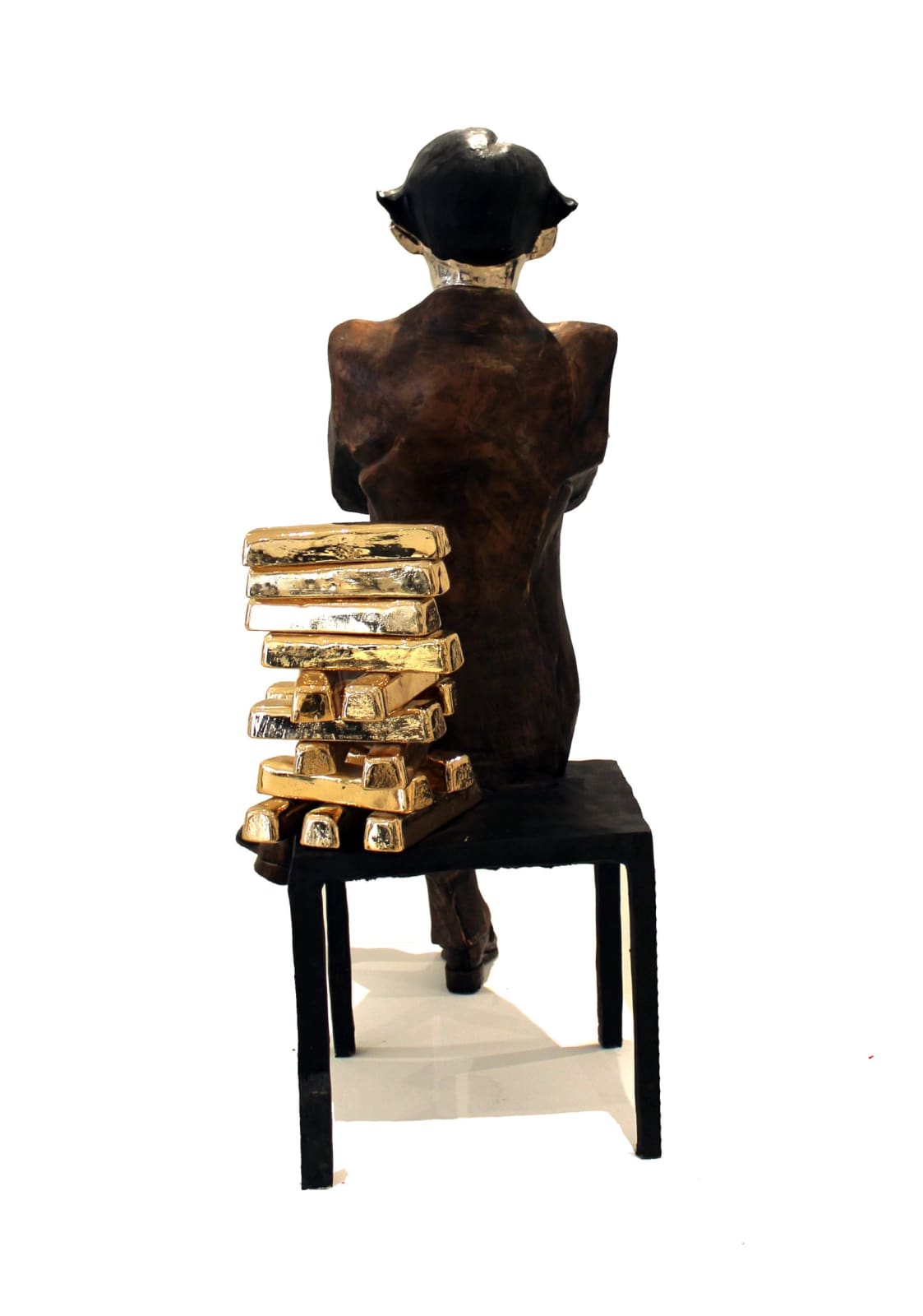 Rear view of a large banker sculpture showing the back of the seated figure. The chair's backrest is made of stacked gold bullion bars, symbolizing wealth, with one chair leg bending under the weight. The sculpture's polished, reflective head contrasts with the textured bronze finish of the suit, highlighting its intricate details.