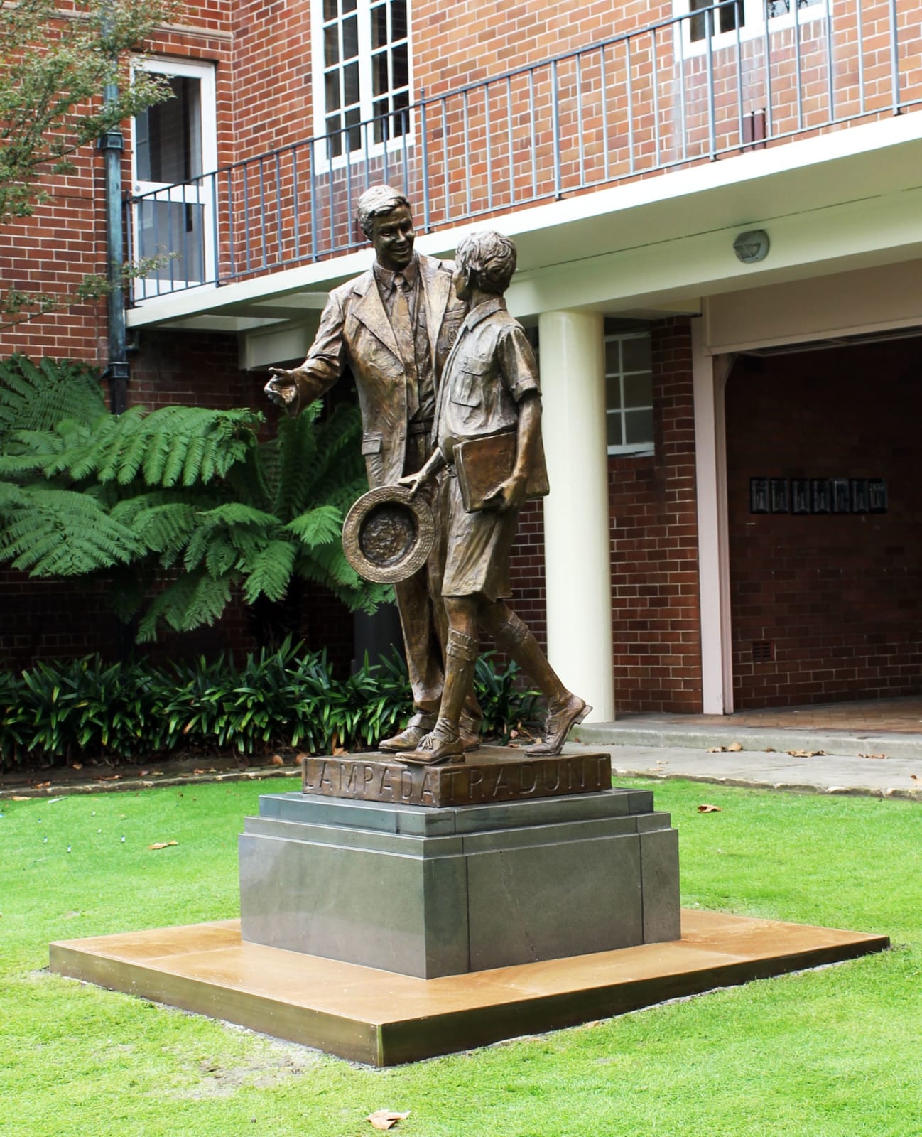 A side perspective of the Shore Boys bronze statue, highlighting the dynamic interaction between the two figures. The setting, with the school's red brick building in the background, complements the statue's theme of education and legacy.