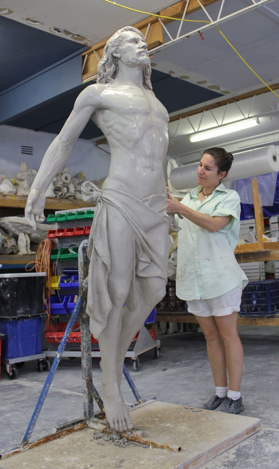 Artist Linda Klarfeld sculpting a life-size clay statue of Jesus ascending. The sculpture features Jesus with outstretched arms and an upward gaze, draped in a flowing cloth around his waist. The scene is set in a studio filled with sculpting tools and materials.
