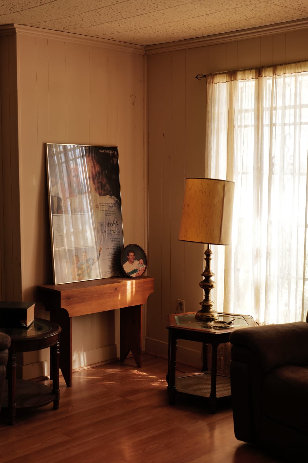 Photograph by Casey Joiner of the corner of a living room setting. It features two side tables, a lamp, and a wooden table against the wall displaying one large rectangular framed image and a small circular one. The sun shines warmly on the sheer white curtains, letting some light reflect of the large image.