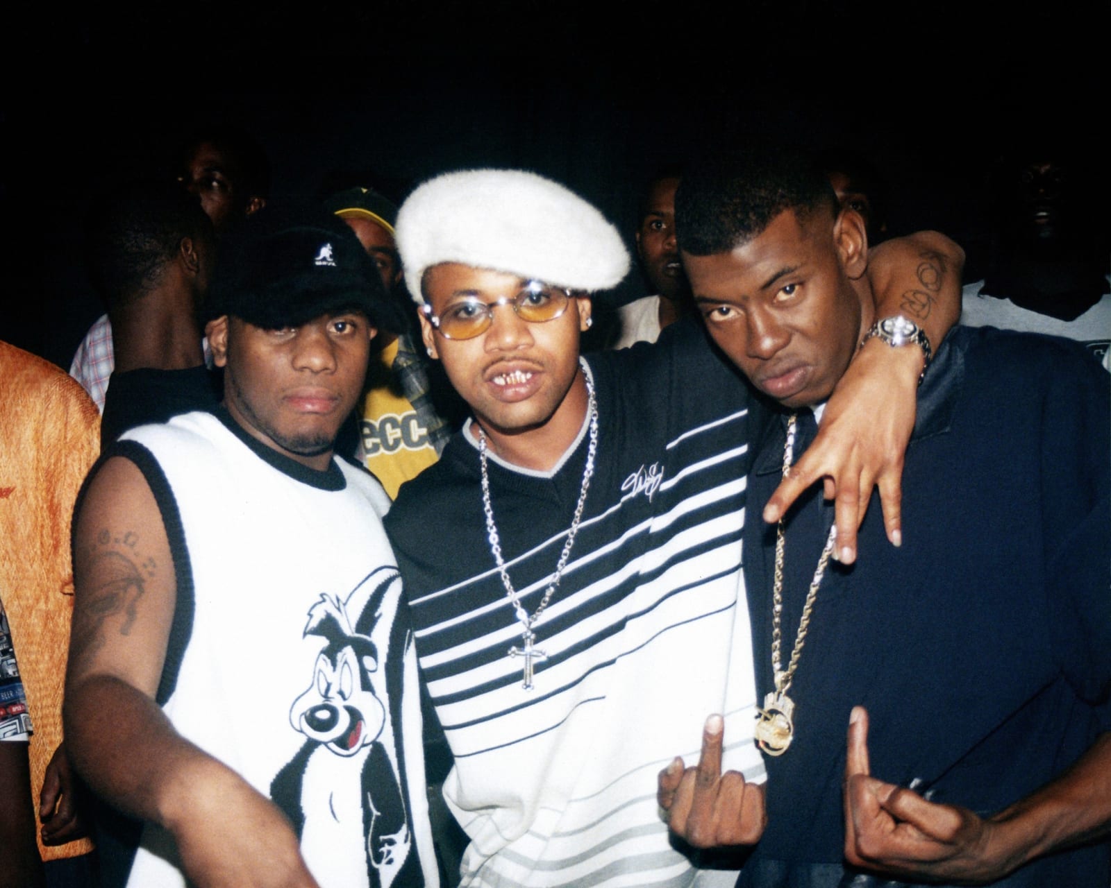 A photo by Polo Silk of rappers 6Shot, Juvenile, and Soulja Slim posing for the camera. 6Shot wears a Kangol baseball cap and a Pepe LePew shirt. Juvenile wears a white fuzzy newsboy cap and yellow tinted glasses. Soulja Slim holds up two middle fingers against his black t-shirt.