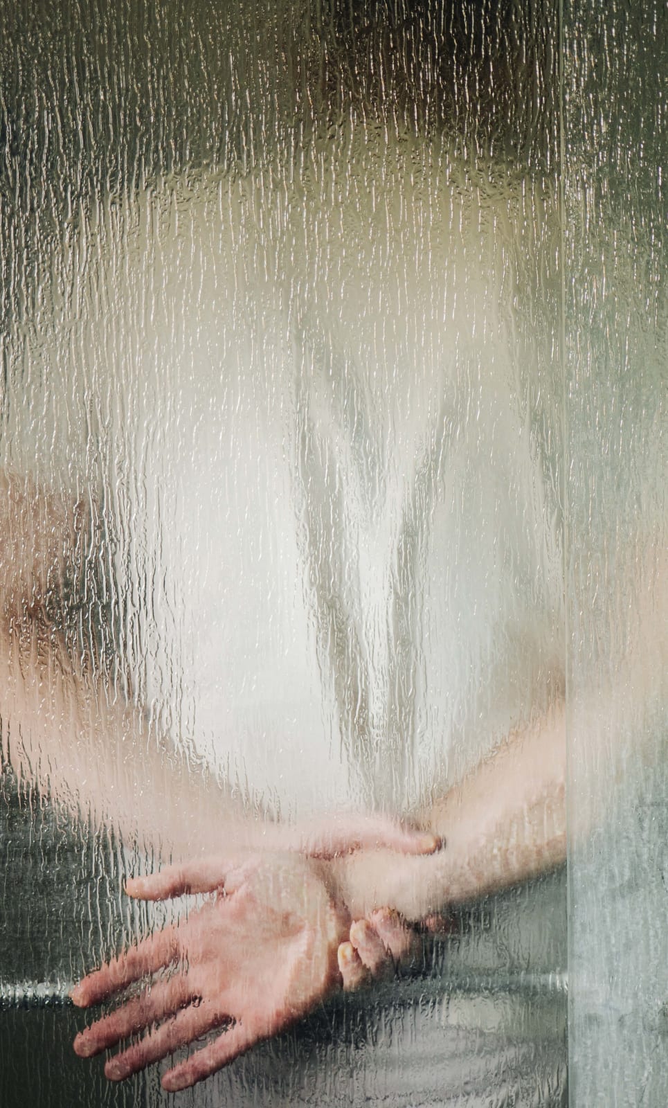 Photograph by Casey Joiner in which the photographer/ viewer looks through a large sheet of vertically textured glass at the back of someone in a white t-shirt who is leaning against the glass with their hands behind their back and one open hand grasped at the wrist by the other.
