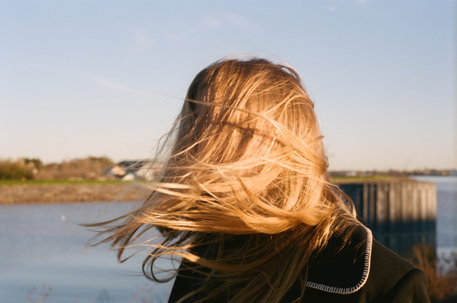 Photograph by Casey Joiner of a woman whose face is obstructed by the wind blowing her hair around. She has blonde hair, rendered brighter still by the evening sun, and she stands in front of the water with it and some land in the background. The sky is an ombre from pale to medium blue.