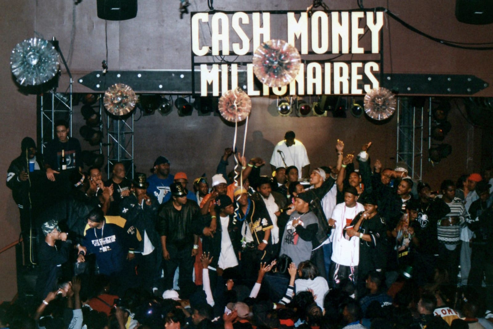 A photograph of members of Cash Money Records altogether on a stage during a party. A sign above them reads 
