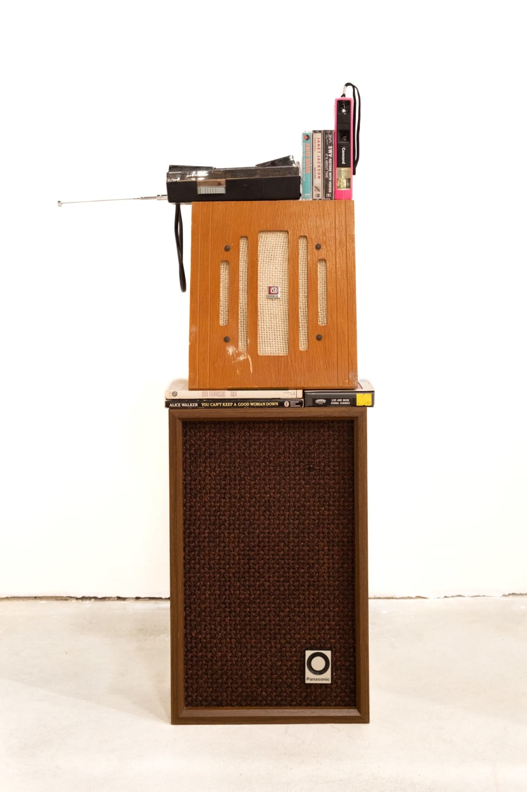 An assemblage sculpture by Jen Everett featuring a vintage speaker, paperback books by Alice Walker and Toni Morrison, two vintage radios, a pink 110 camera, and casette tapes by Donna Summers, Janet Jackson, Anita Baker, and SWV